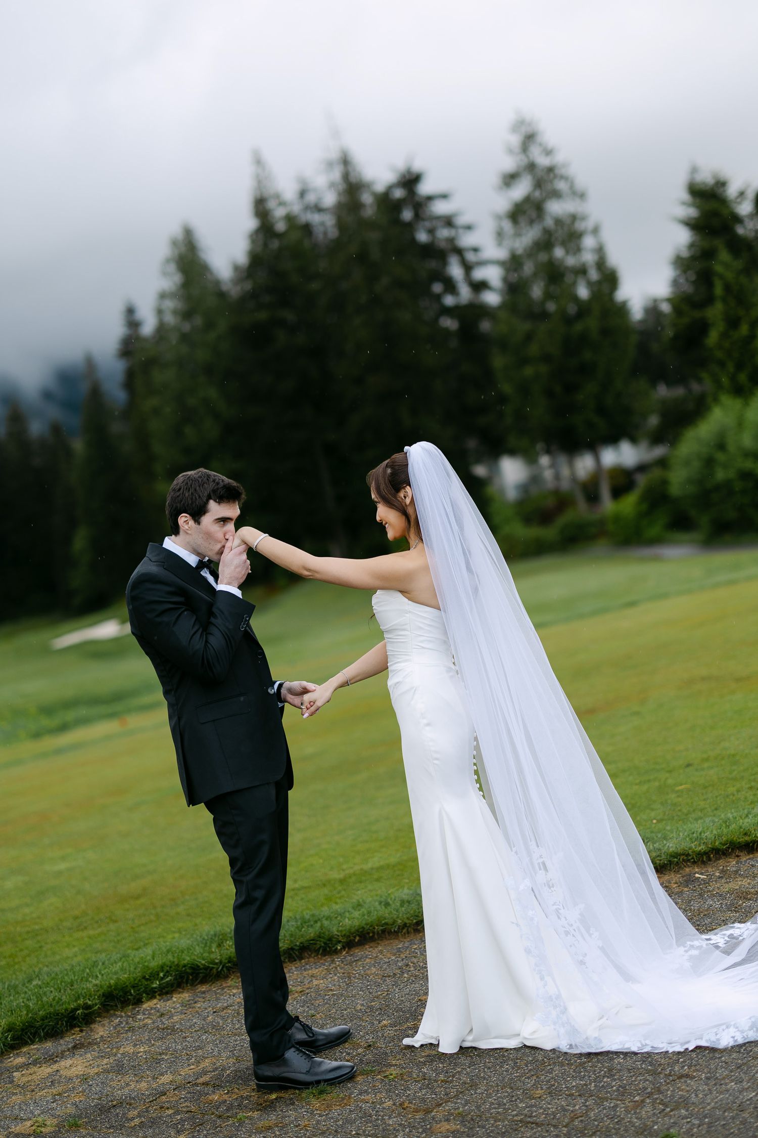 Groom in black tuxedo kisses bride's hand while she holds her white gown and veil on a lush green golf course.