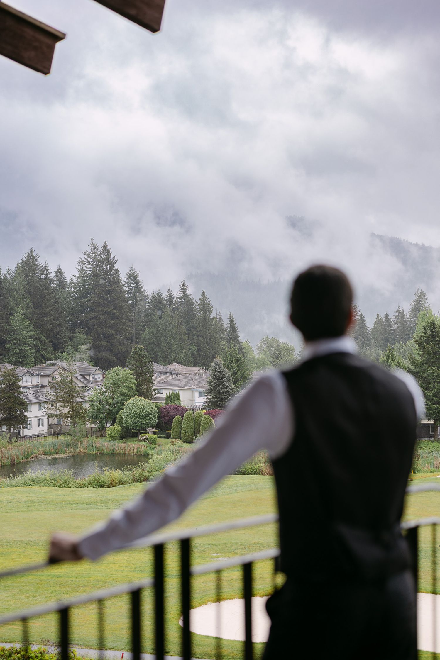 A groom in a vest stands on a balcony overlooking a misty mountain landscape with evergreen trees and a pond.