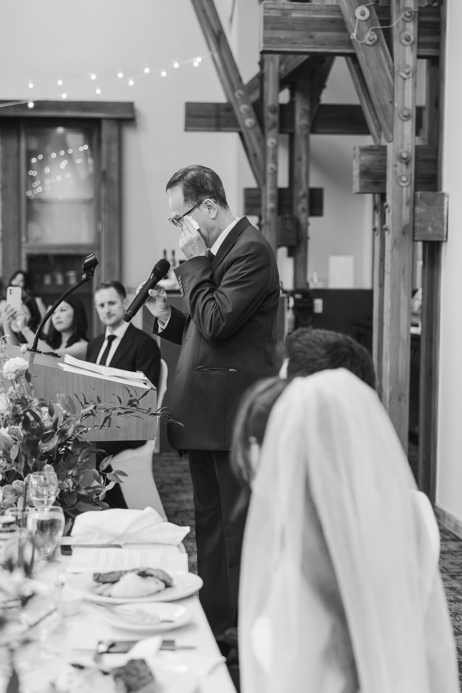 Emotional man in suit giving tearful wedding speech at podium beside decorated banquet table.