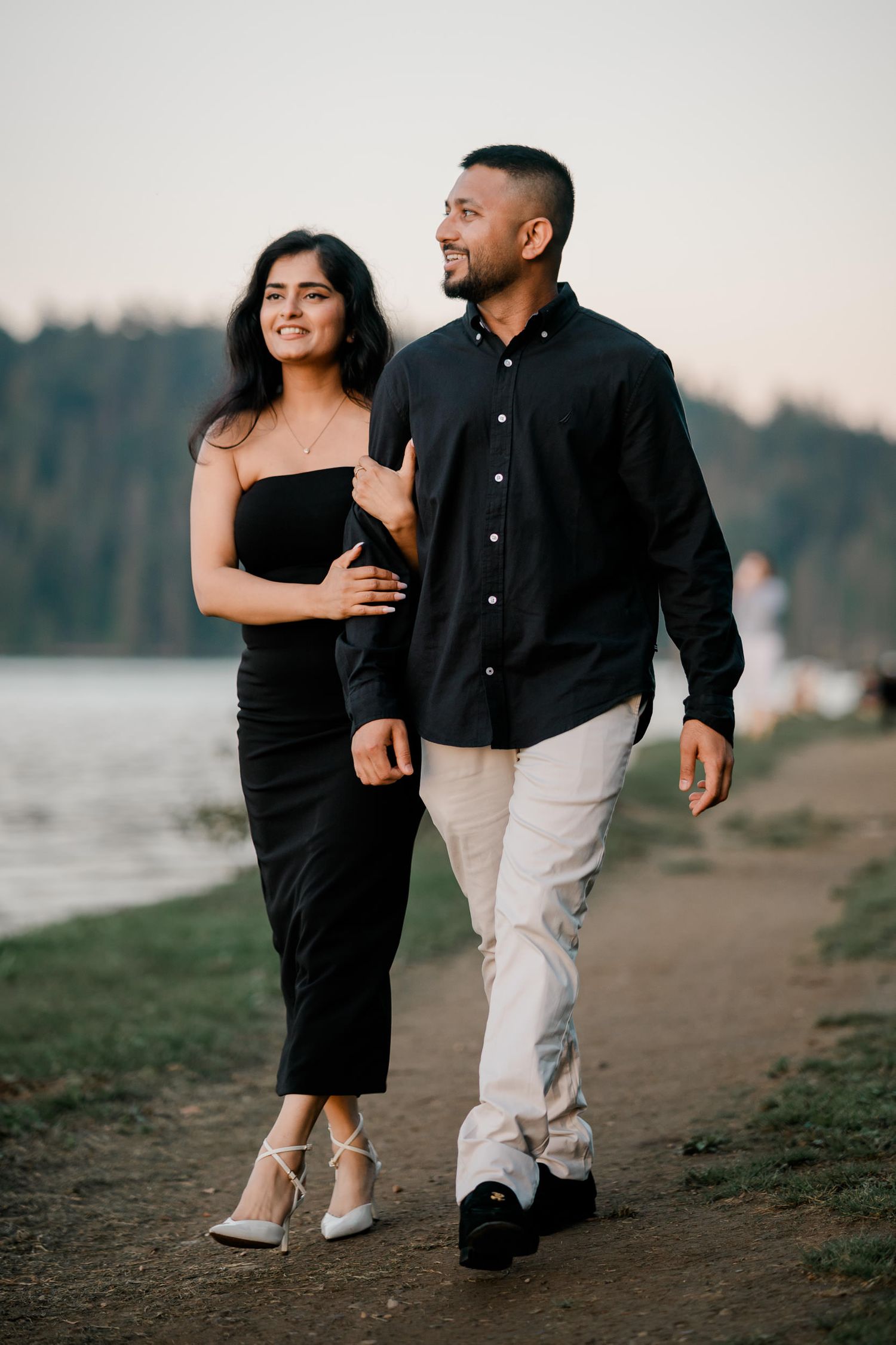 A couple in black and beige outfits walking together on a beach path at sunset.