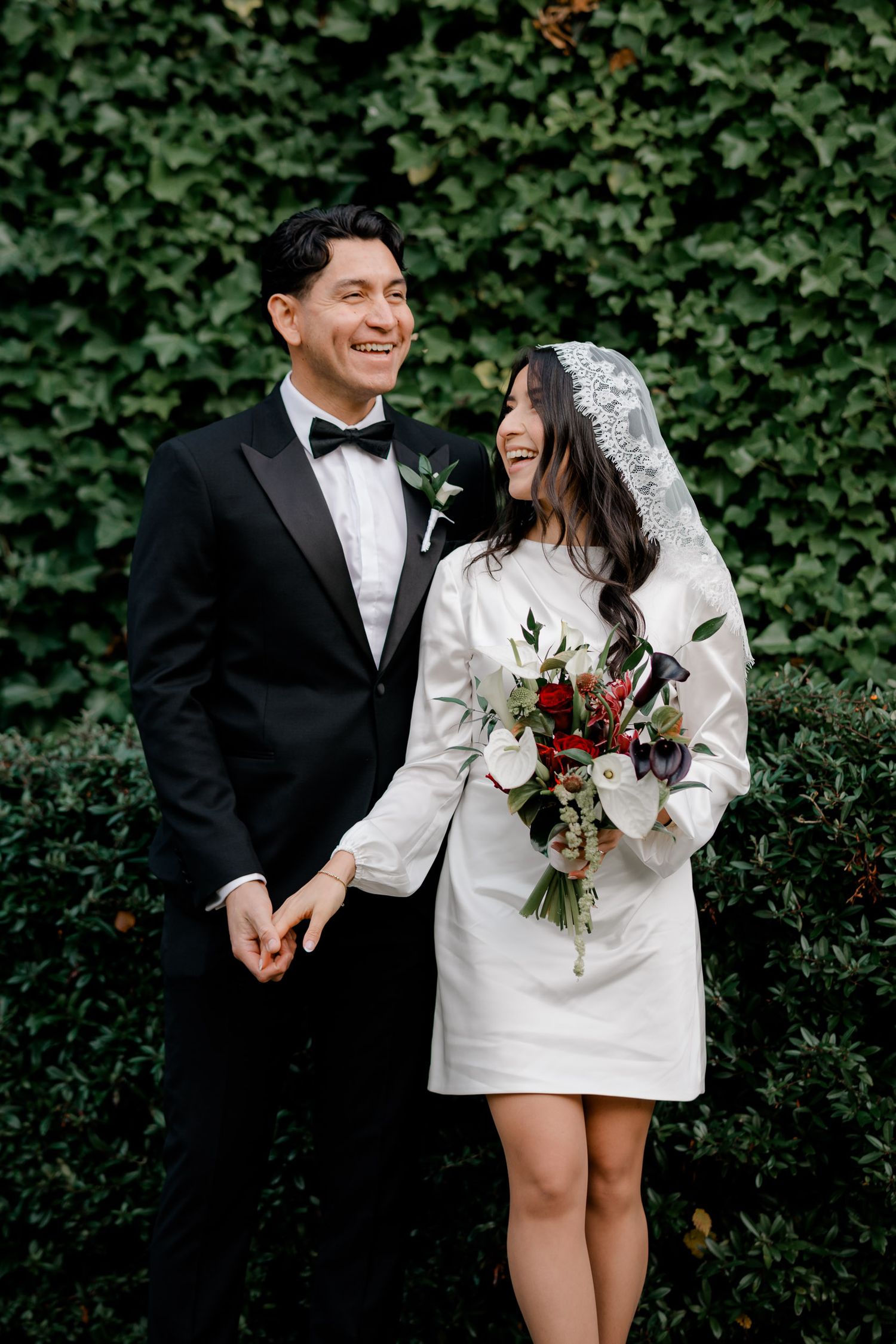 A joyful couple shares a laugh during their wedding day against a lush green ivy wall backdrop.