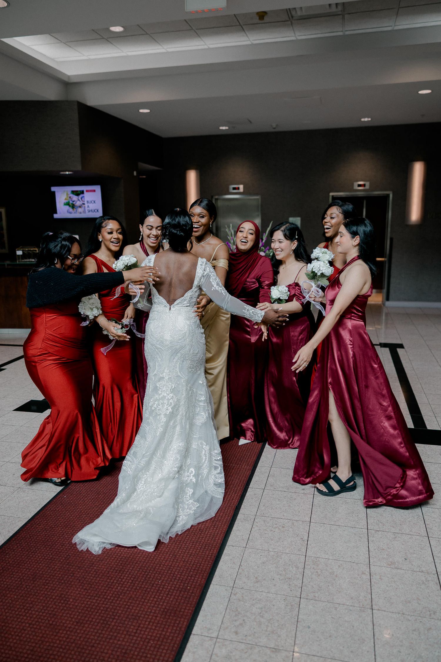 Bride in white lace gown poses playfully with bridesmaids wearing burgundy dresses on tiled floor at wedding reception.