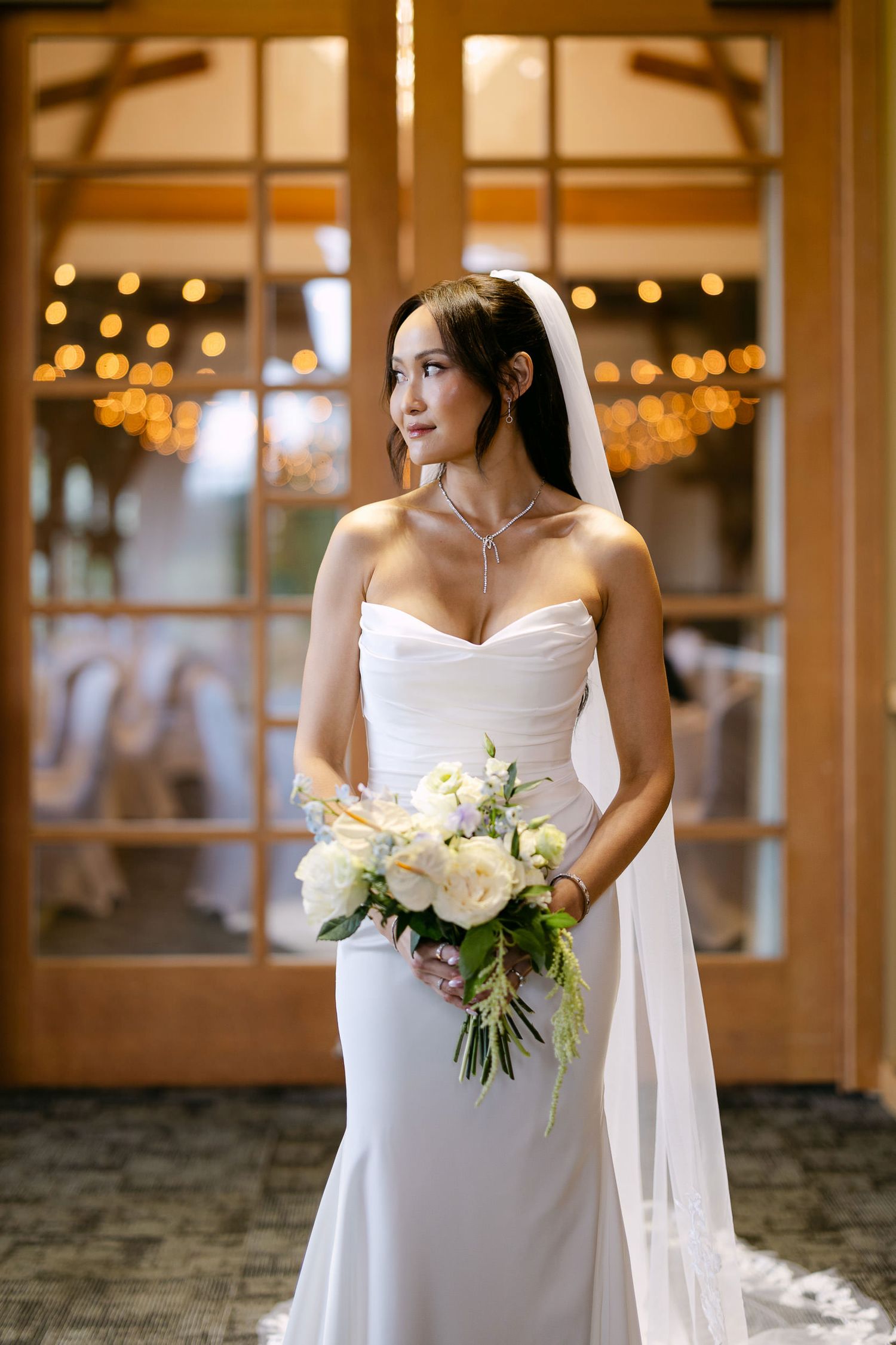 Bride in strapless white gown and veil holding white floral bouquet standing before warmly lit venue doors.