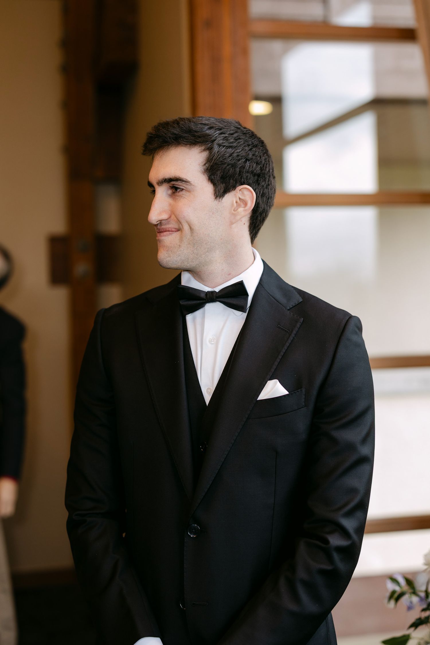 A groom in a classic black tuxedo with bow tie and white pocket square smiles warmly during his wedding ceremony.