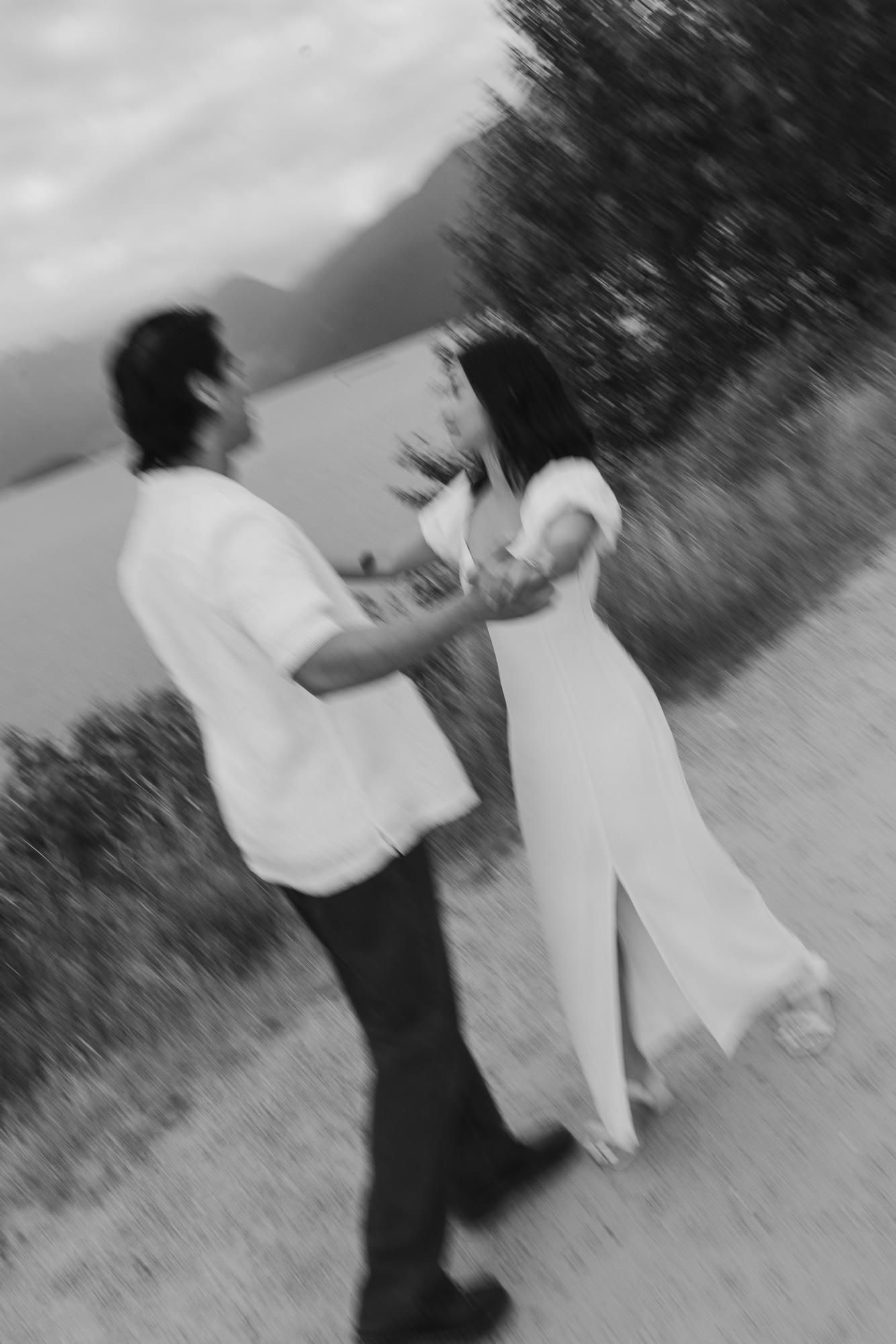 Motion-blurred black and white photo of a couple dancing hand in hand beside a lake with trees and mountains.