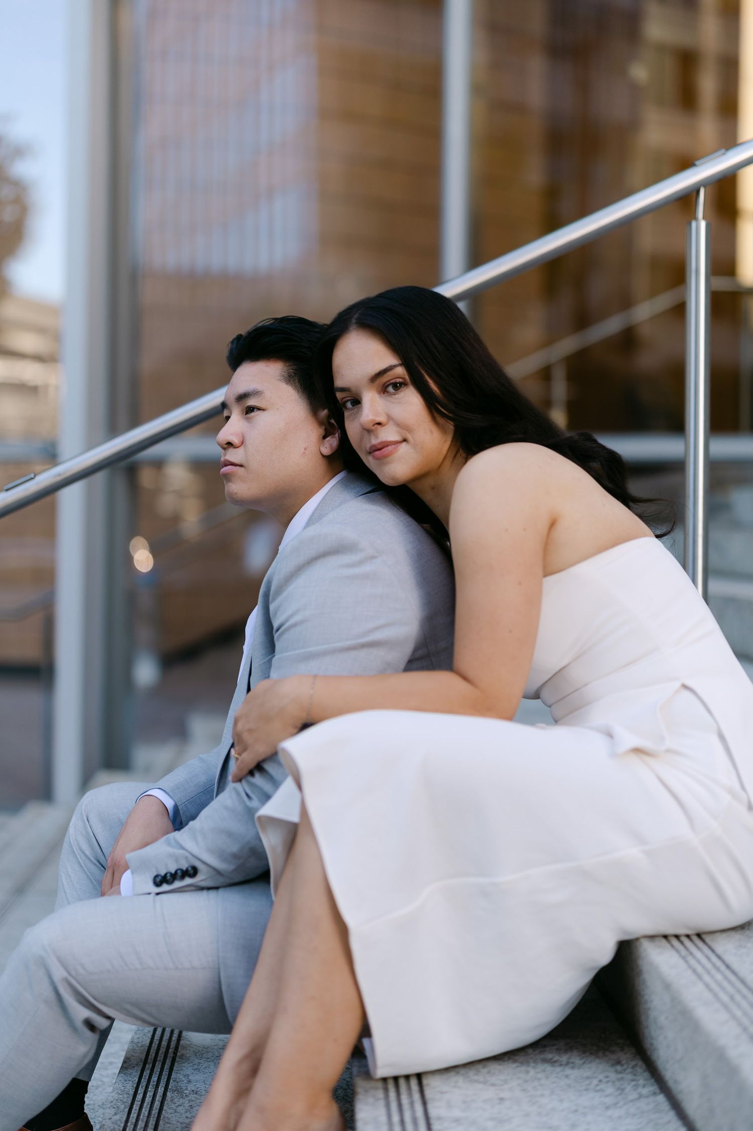 Couple in a white dress and light gray suit sit close on outdoor city steps as she leans her head against his shoulder.