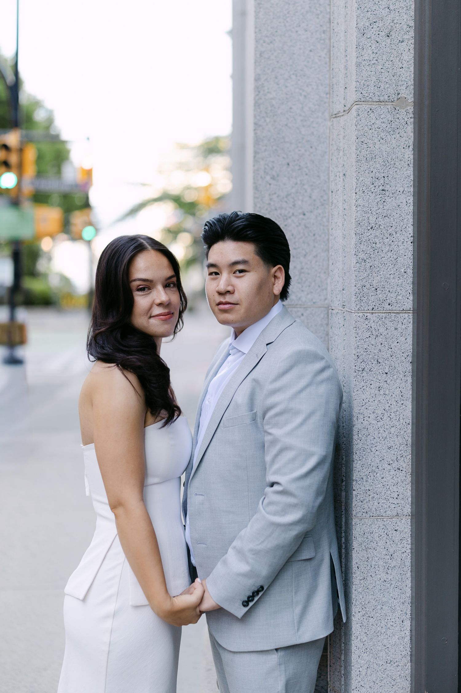 Engagement portrait of a couple holding hands against a stone wall, with city traffic lights softly blurred behind them.