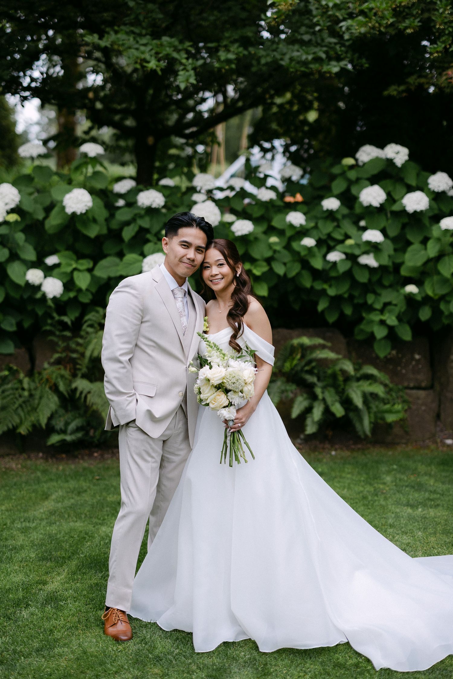 Bride and groom pose together in a lush garden with white hydrangeas, she holds a white floral bouquet in a ball gown.