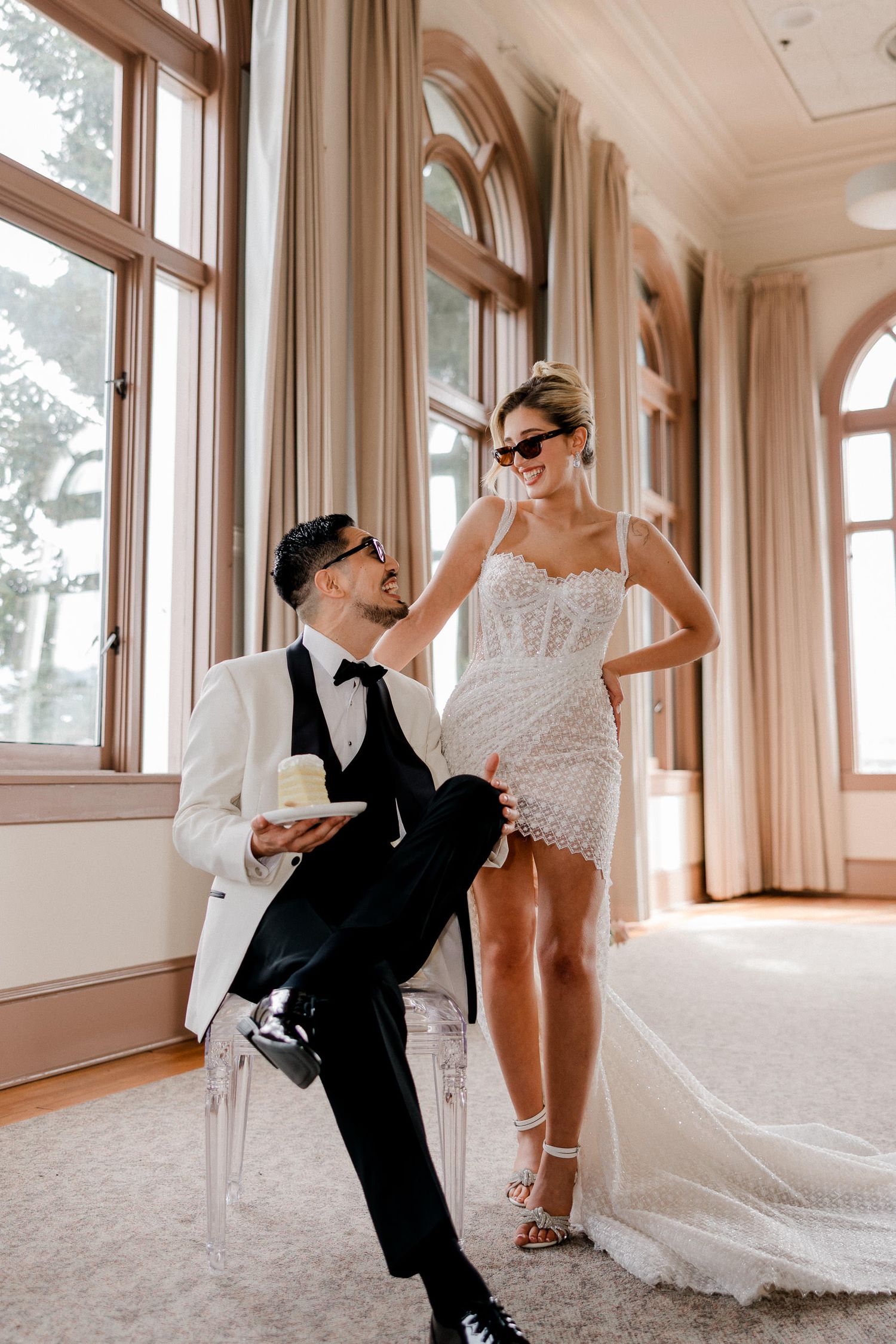 A playful wedding photo series showing a couple dancing and posing in a sunlit hallway with large windows.