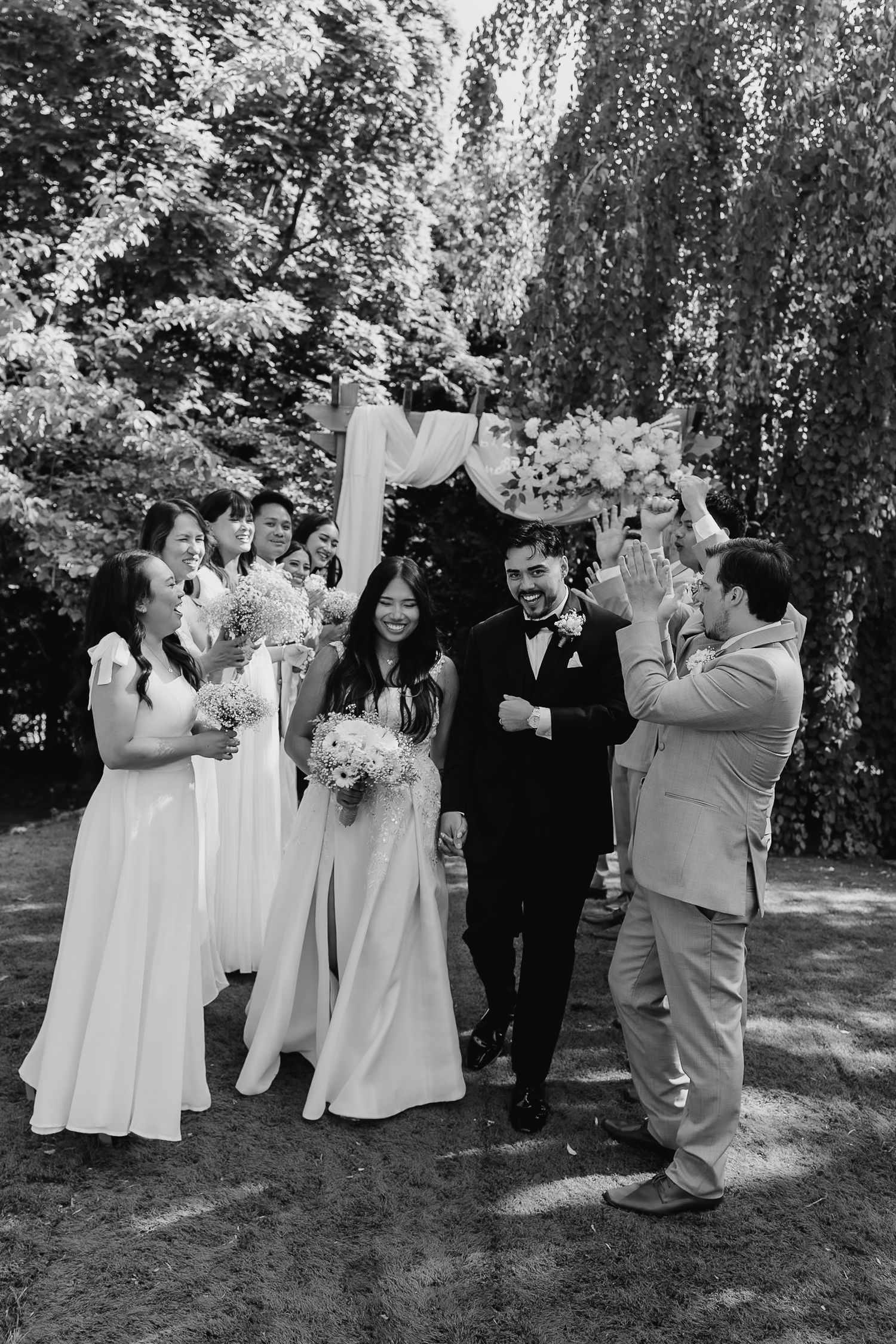A black and white wedding ceremony photo showing a celebration under trees with attendants in formal attire.