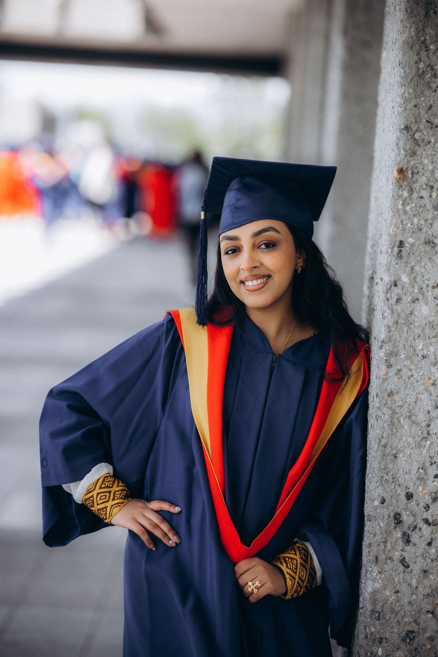 Graduate in navy blue cap and gown with orange and gold accents poses for graduation photos against concrete columns.