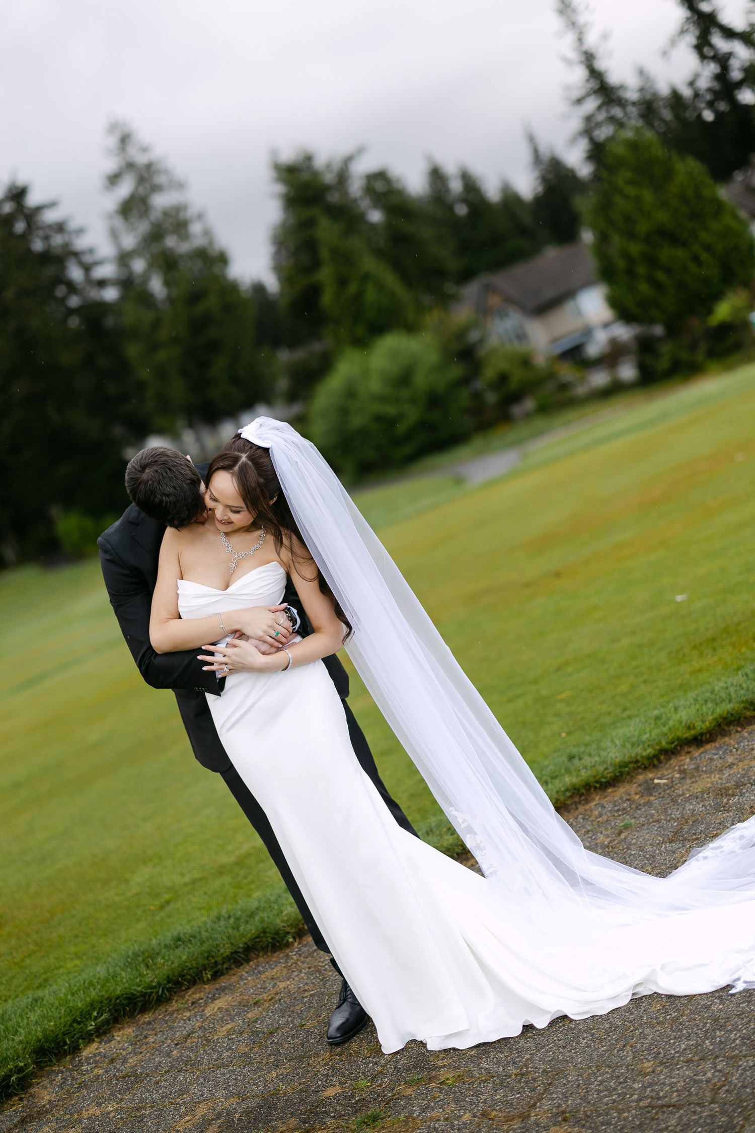 Bride and groom embrace on a lush green lawn, her long veil trailing behind her.