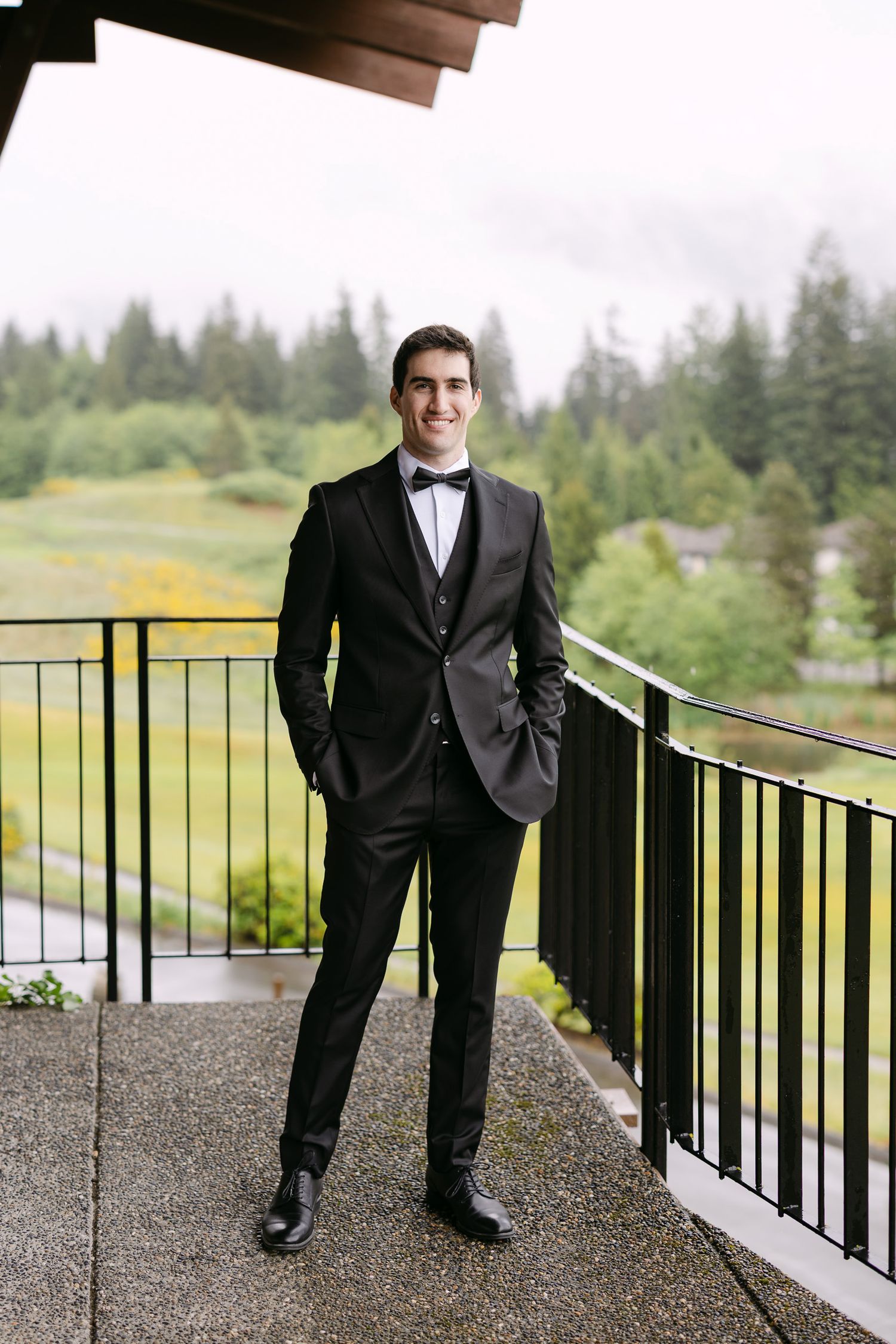Groom in black tuxedo with bow tie stands on outdoor balcony with lush green forest backdrop.