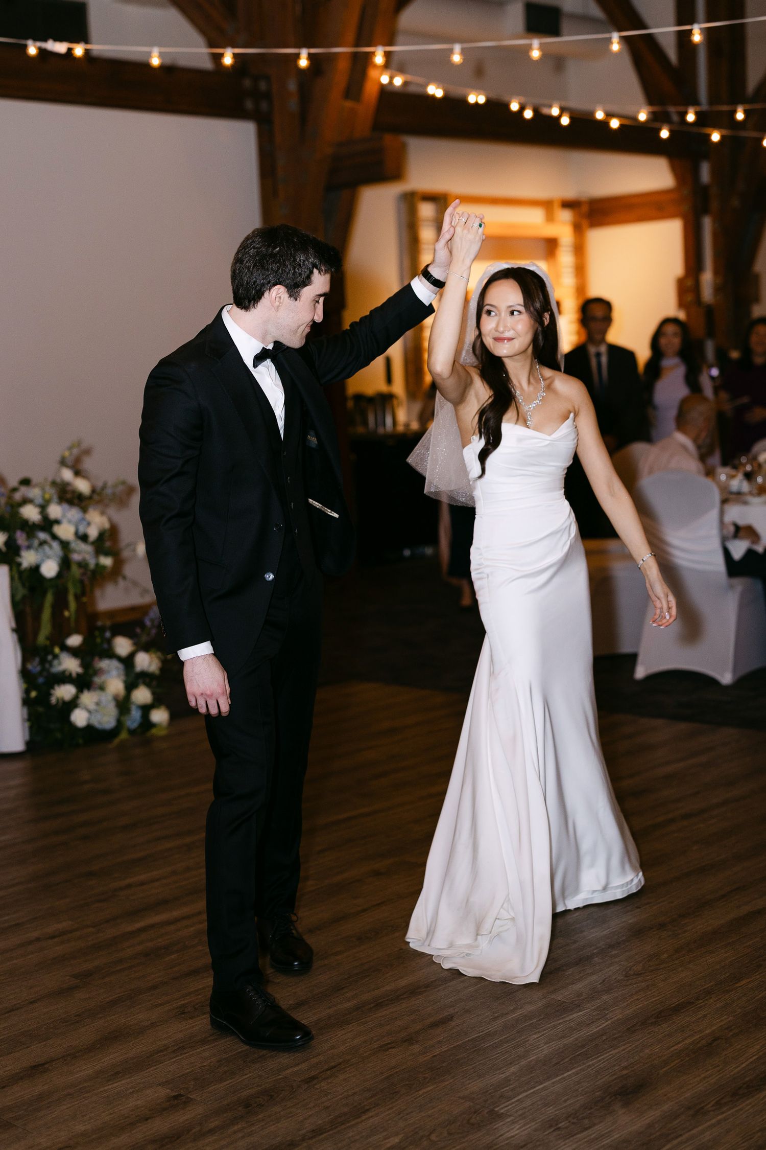 Bride in white gown and groom in black tuxedo share first dance under string lights at elegant wedding reception.