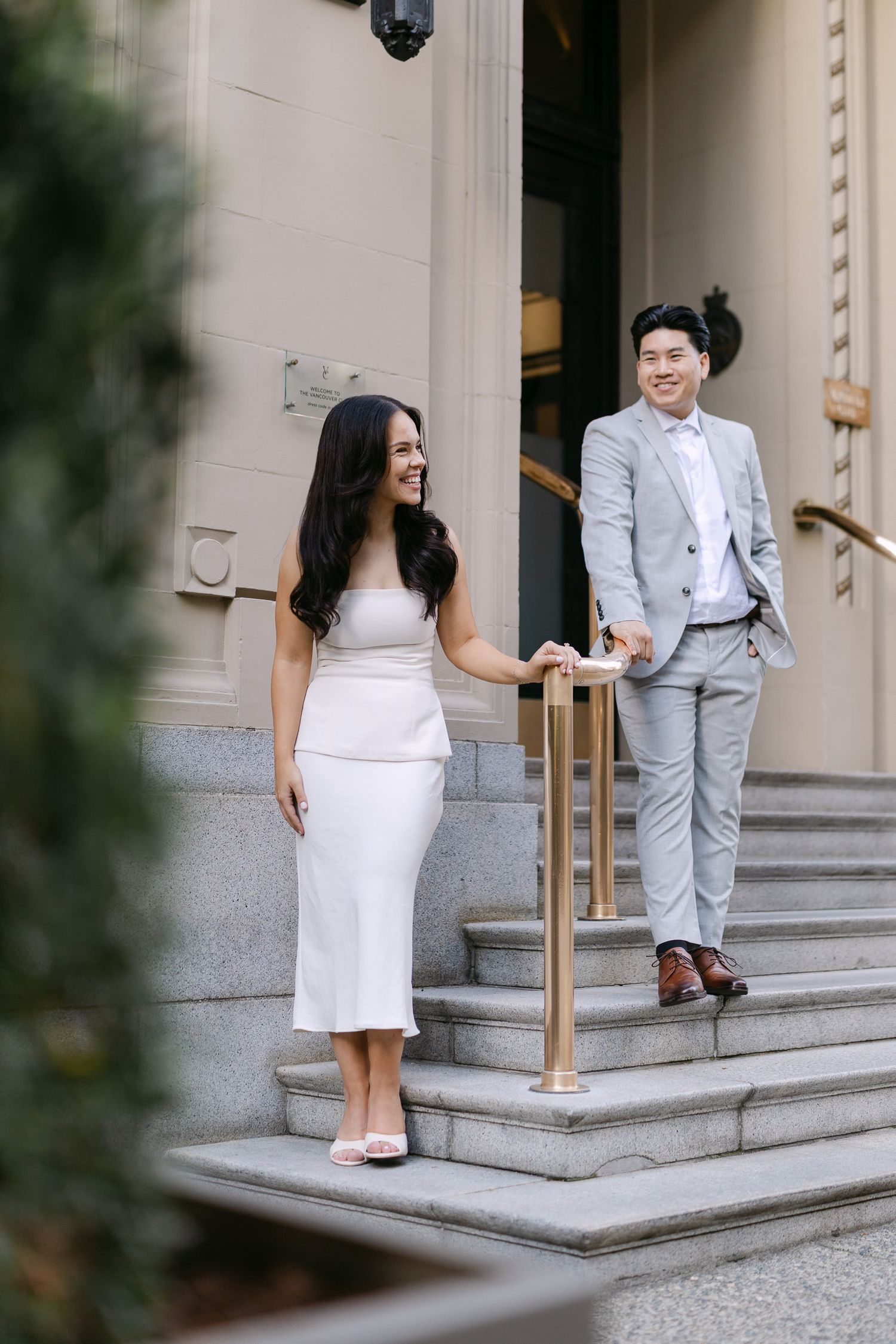 Smiling couple in white dress and light gray suit pose on stone steps with a gold railing during an engagement photo session.