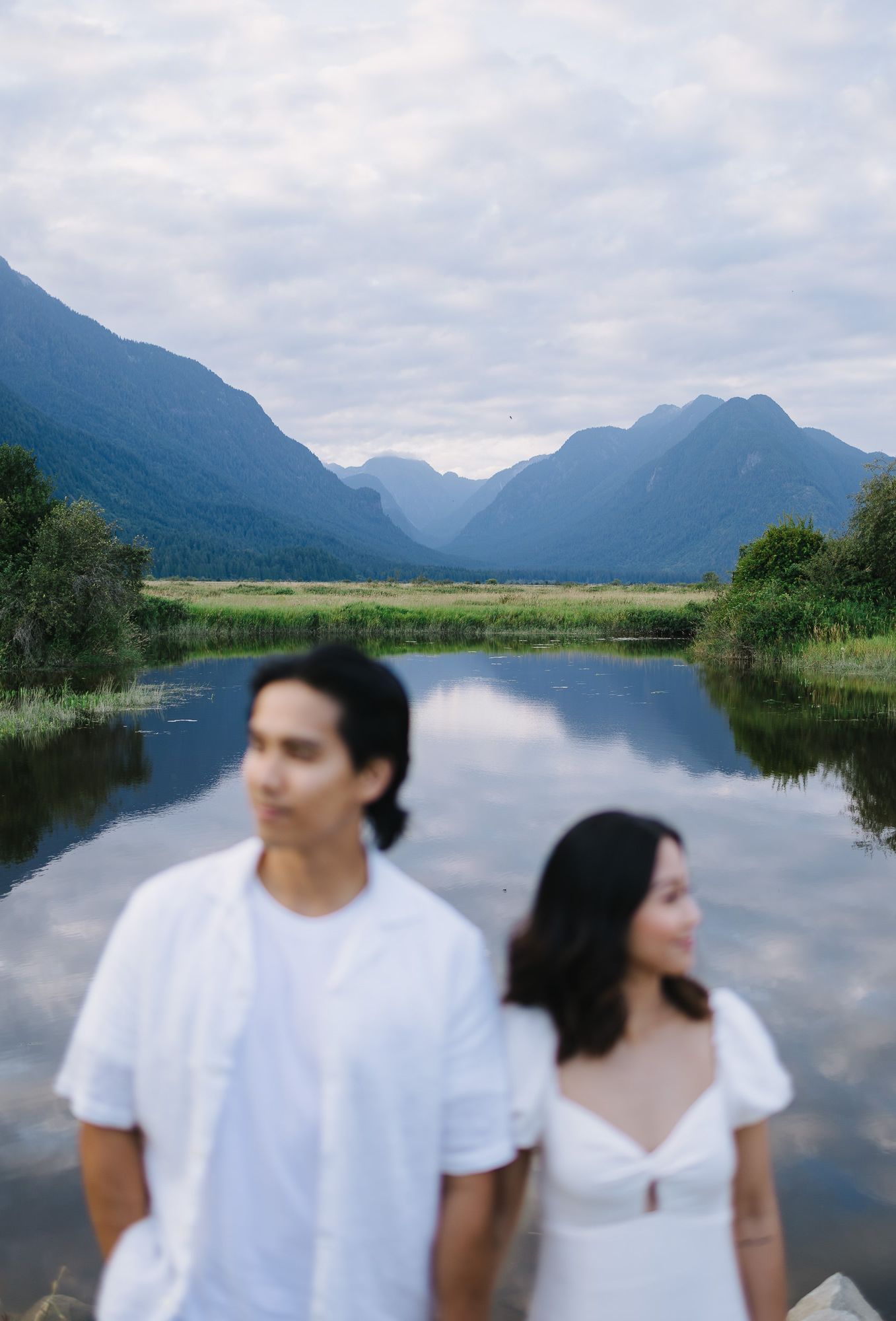Couple in white outfits standing beside a calm reflective lake with misty mountains and cloudy sky in the background.