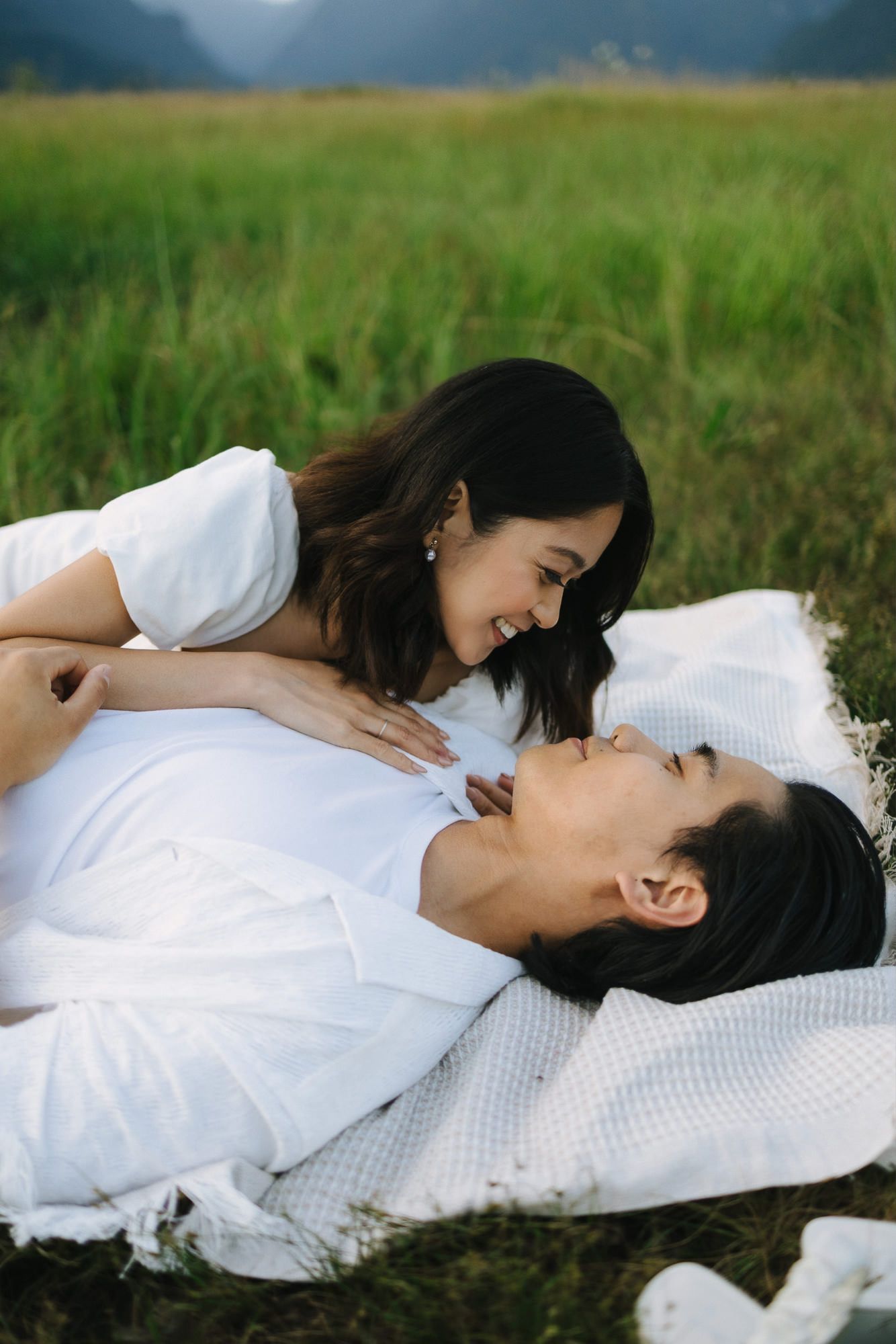 Smiling couple in white outfits lie close together on a blanket in a green meadow with mountains behind.