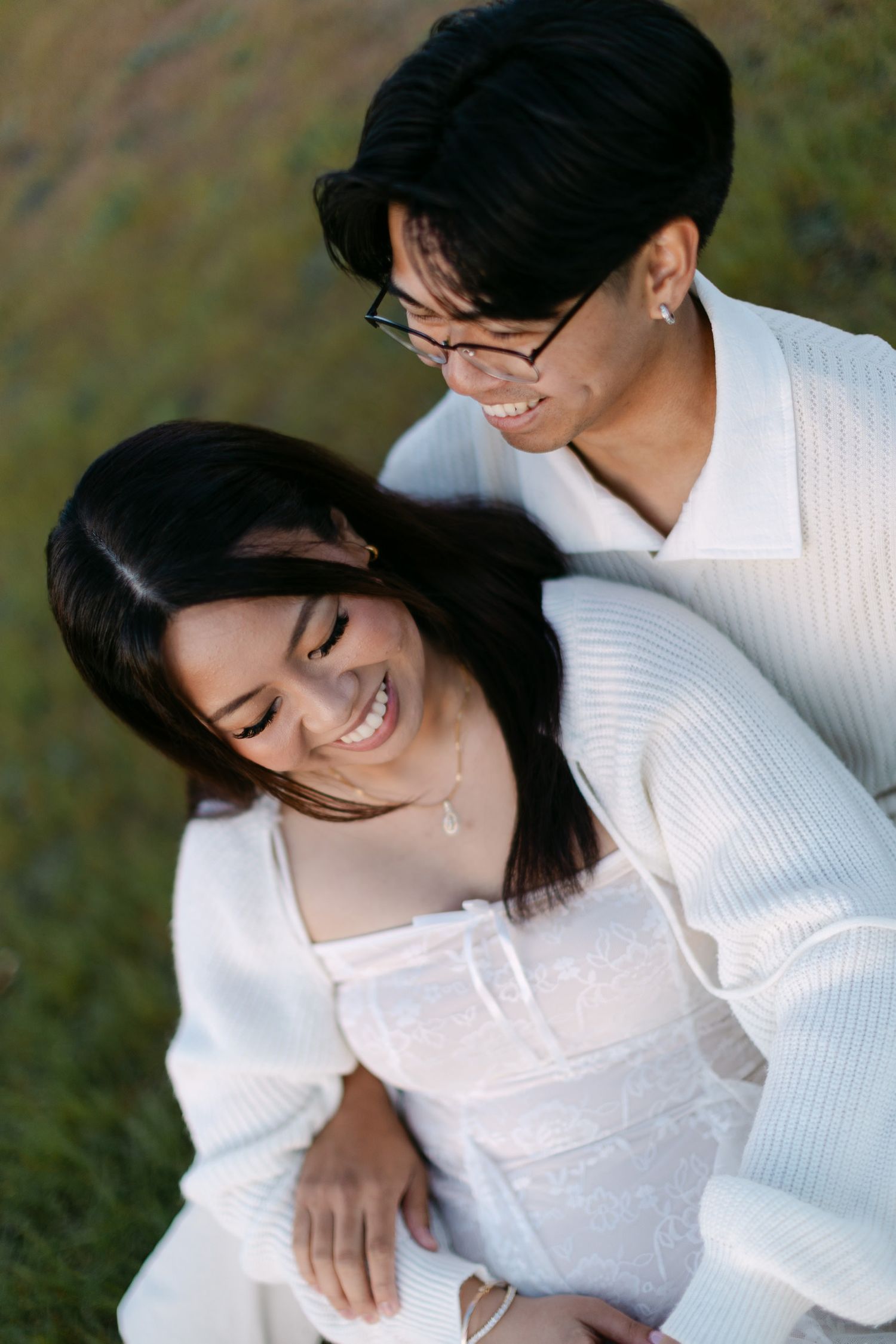 A smiling couple in white outfits embraces on a grassy hillside, the woman laughing as the man smiles down at her.