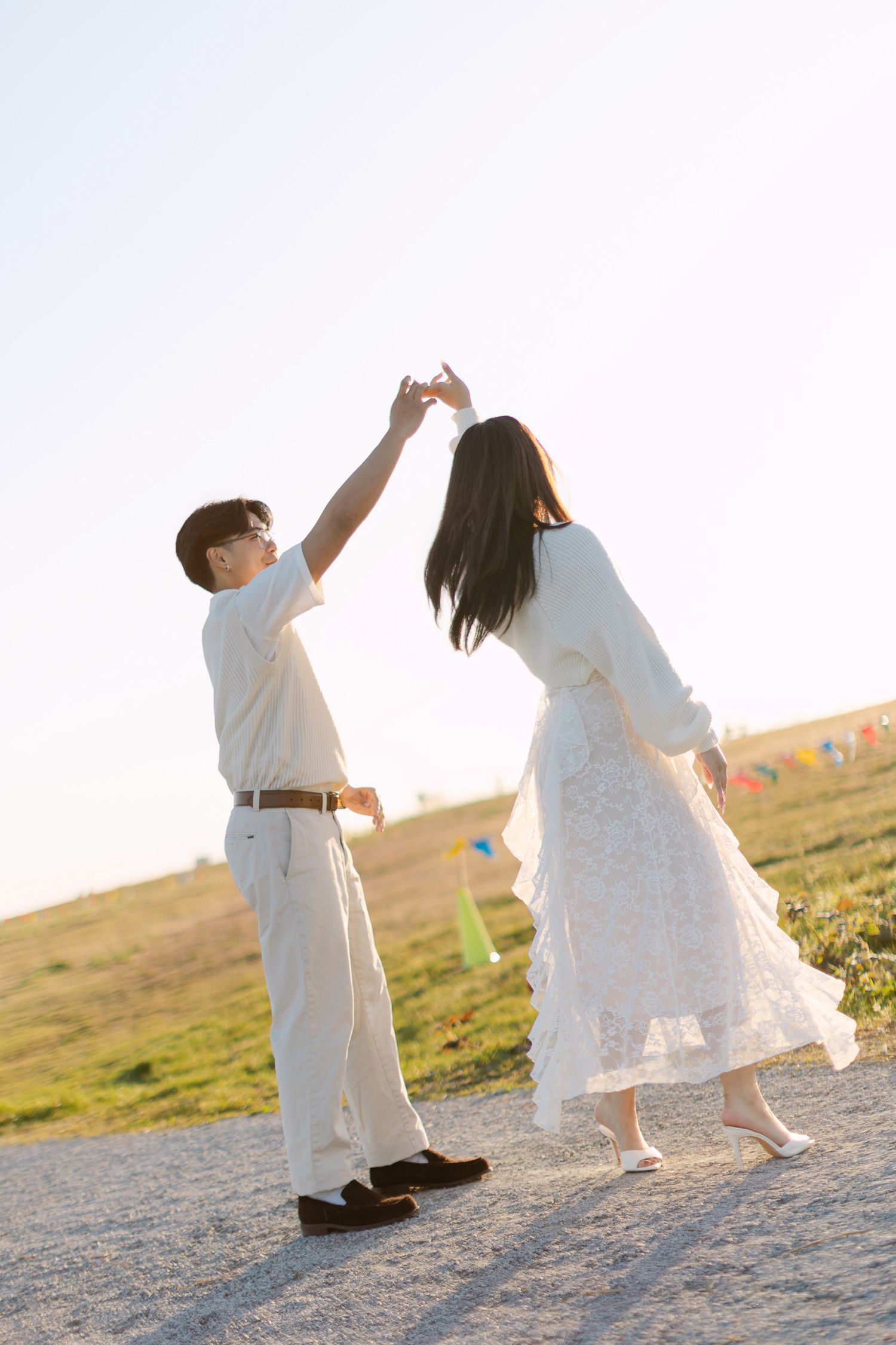 Couple in white outfits dancing and twirling on a gravel path near a grassy hill at golden hour during an engagement photo session.
