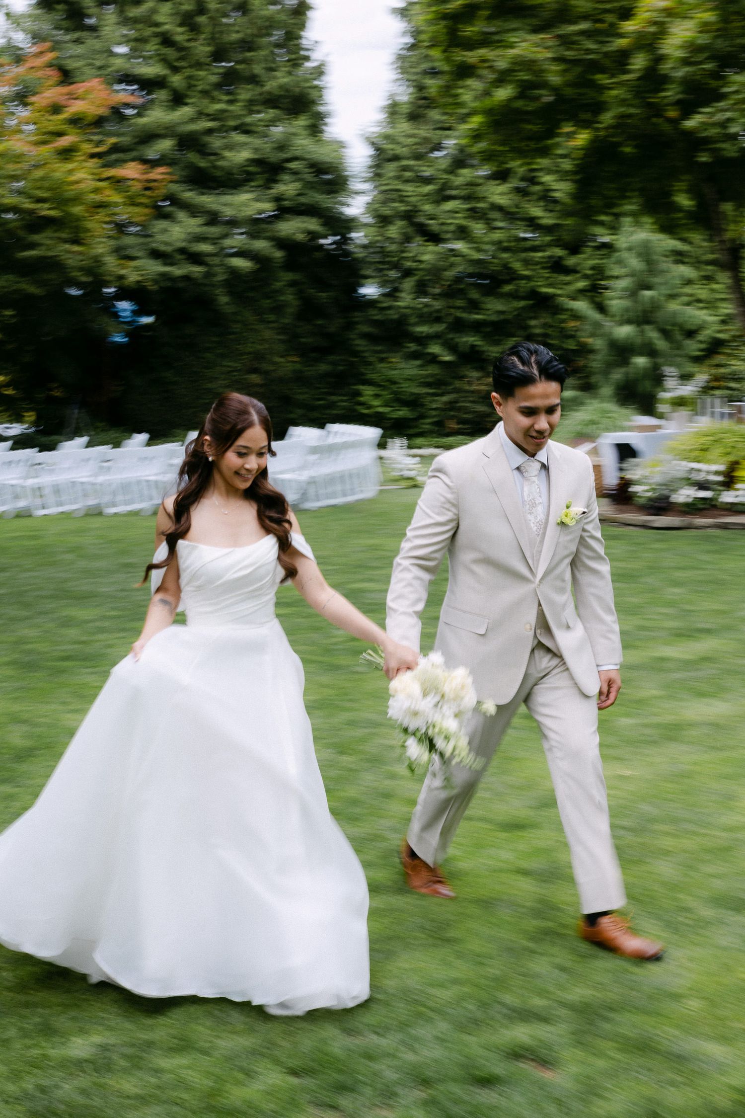 Bride and groom walking hand in hand across green lawn at outdoor wedding ceremony with white chairs.
