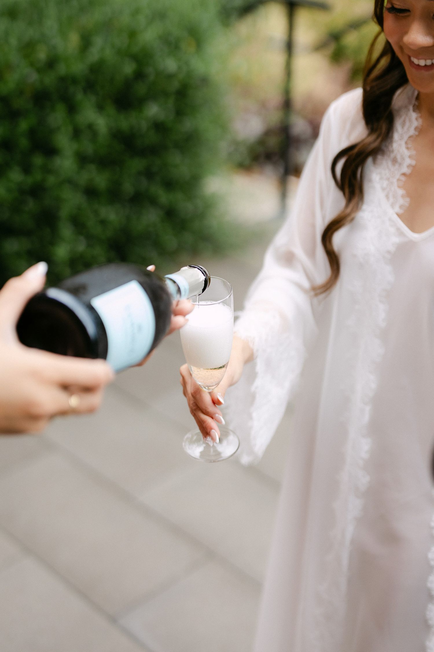 Bride in white robe holding champagne glass being filled at outdoor wedding.