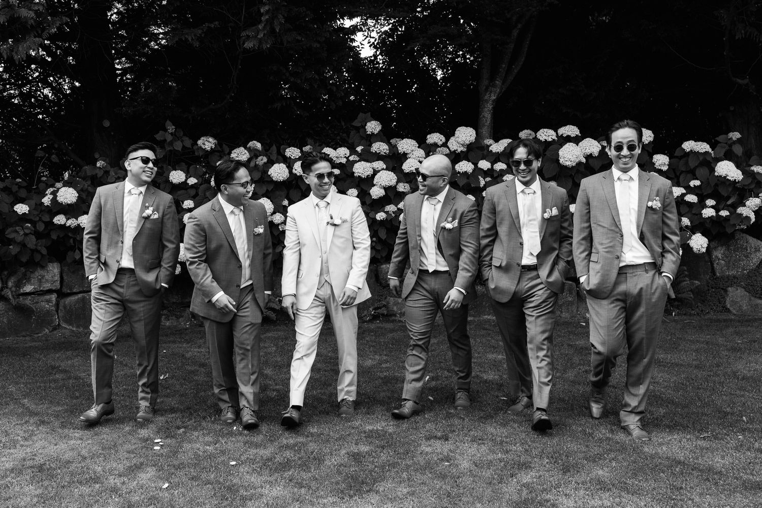 Black and white photo of groom in white suit with groomsmen in gray suits posing outdoors before hydrangea bushes.