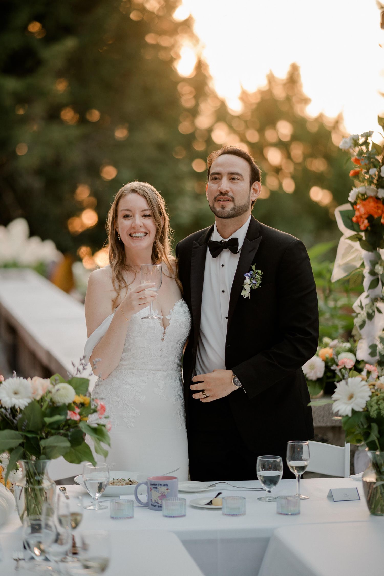 A couple in wedding attire stands at their reception table during golden hour with floral decorations and glassware.