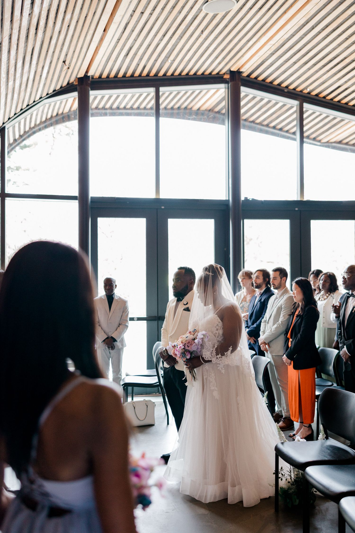 A group of wedding guests gathered in a modern venue with wood slat ceiling and large windows.