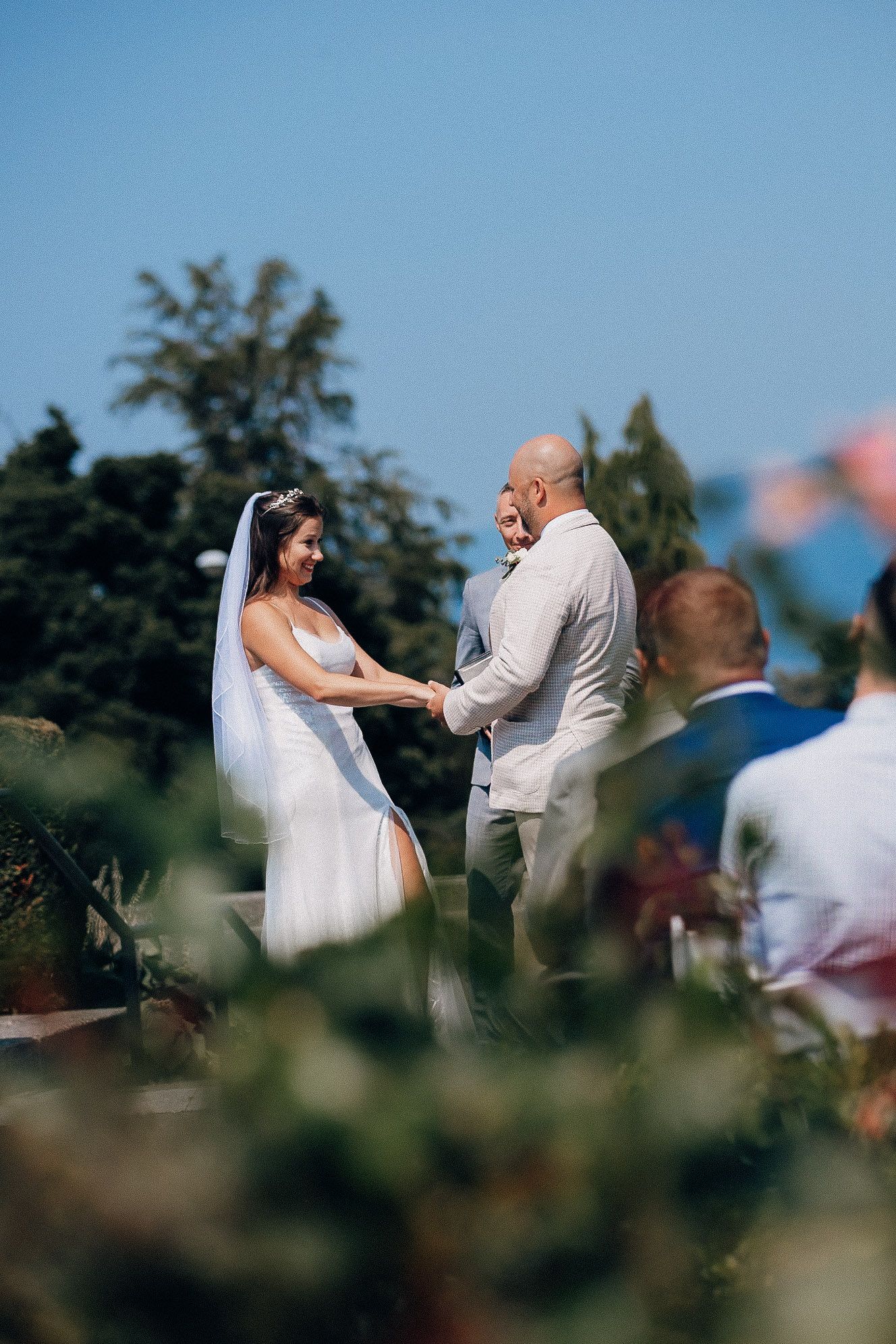 A couple holds hands during an outdoor wedding ceremony surrounded by greenery on a sunny day.