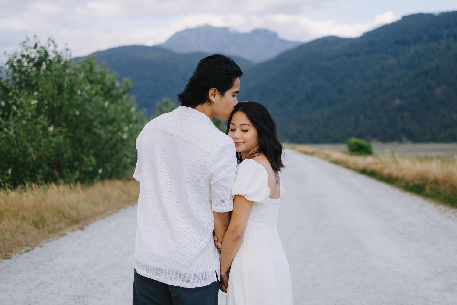 A couple in white clothing embraces on a mountain path surrounded by scenic natural landscape and rolling hills.