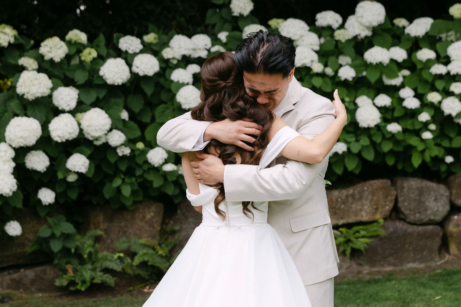 Bride and groom share a heartfelt embrace in front of lush white hydrangea bushes at an outdoor wedding.