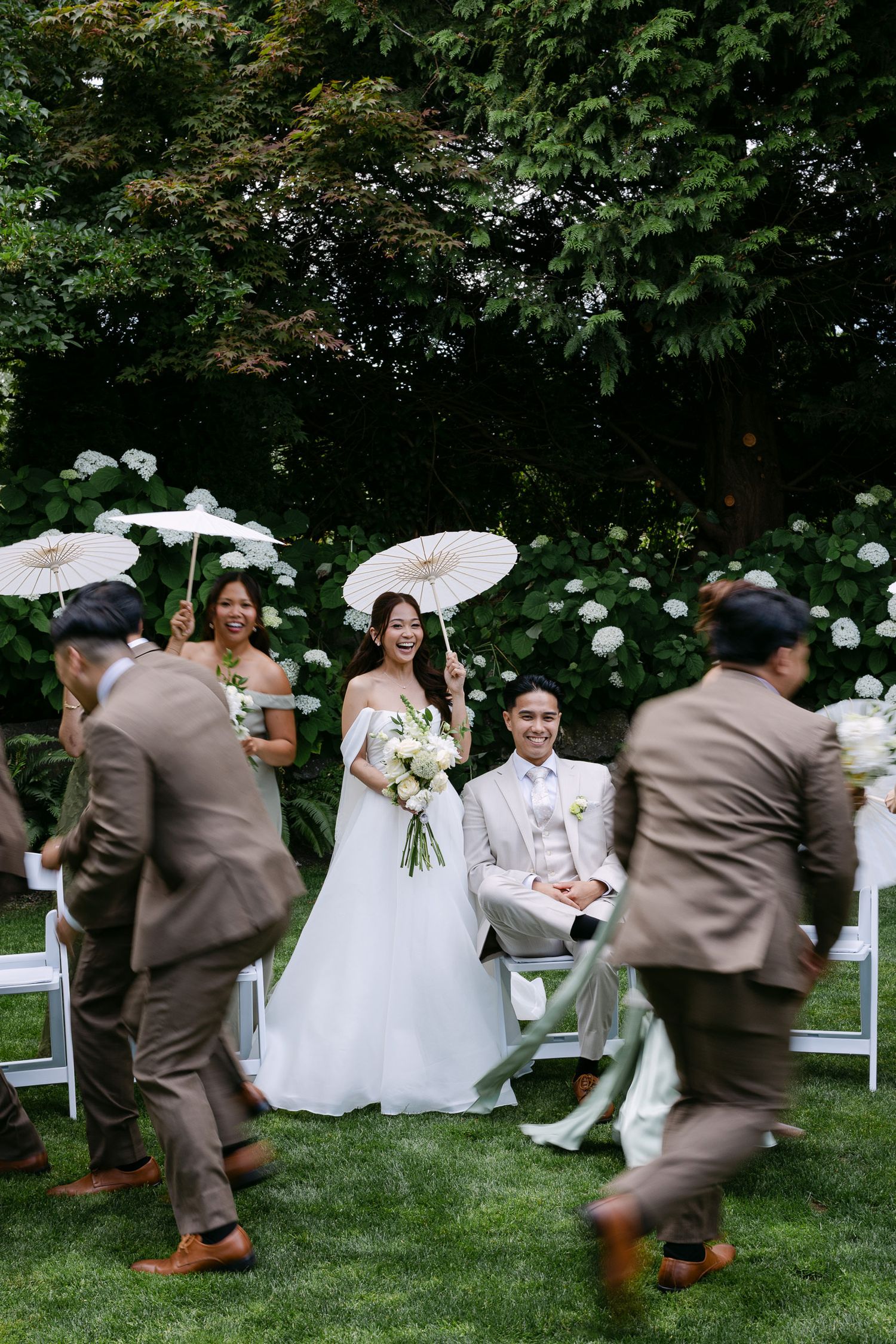 Bride and groom smile during outdoor garden wedding ceremony with bridesmaids holding white parasols and groomsmen in brown suits.
