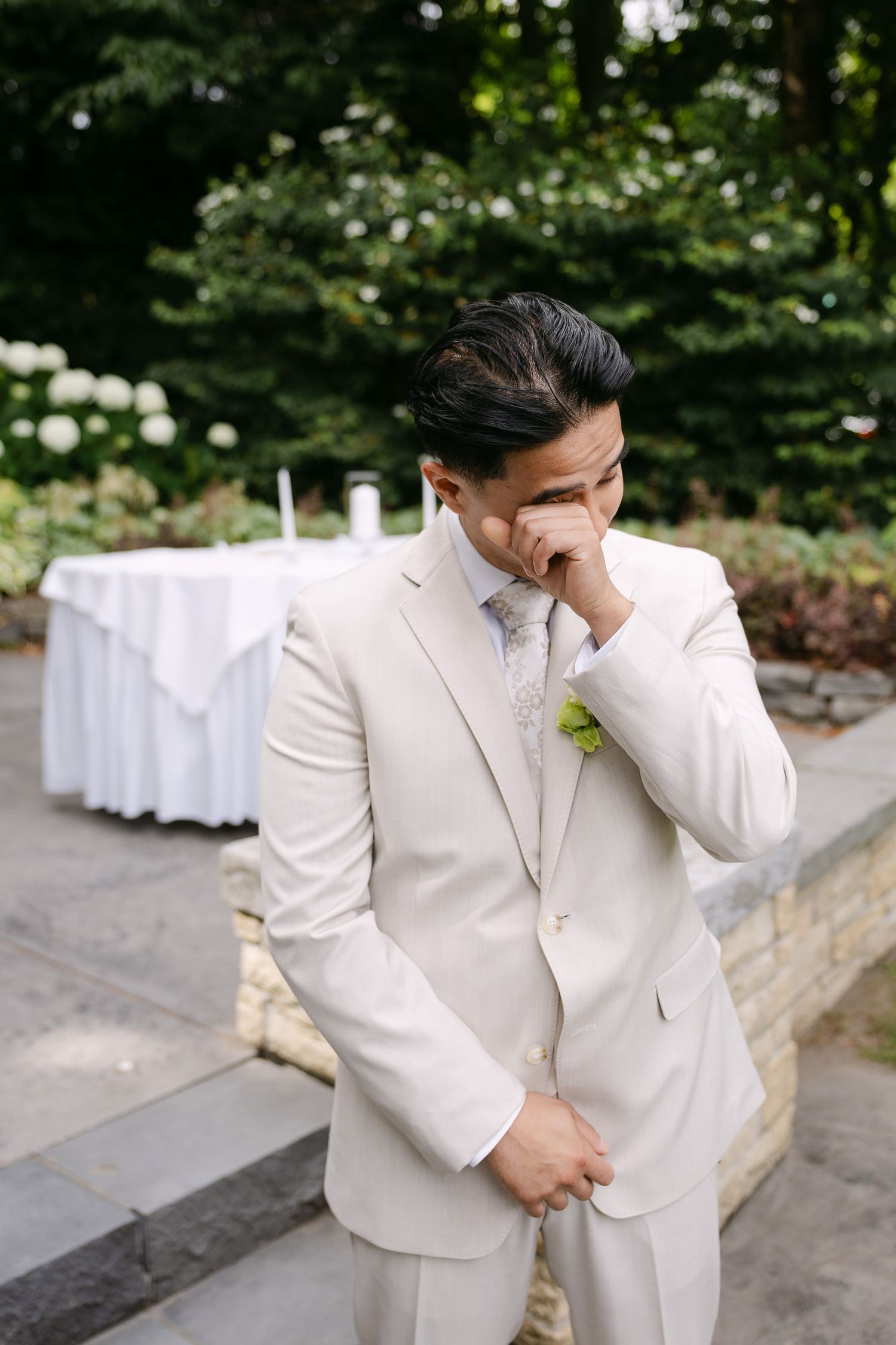 Emotional groom in cream suit wipes tears during outdoor wedding ceremony surrounded by greenery.