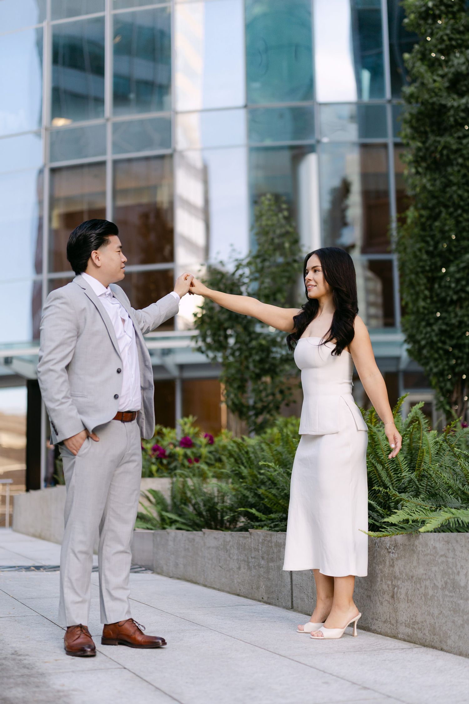 Woman in a white dress twirls with her partner in a gray suit on a downtown sidewalk in front of a glass building.