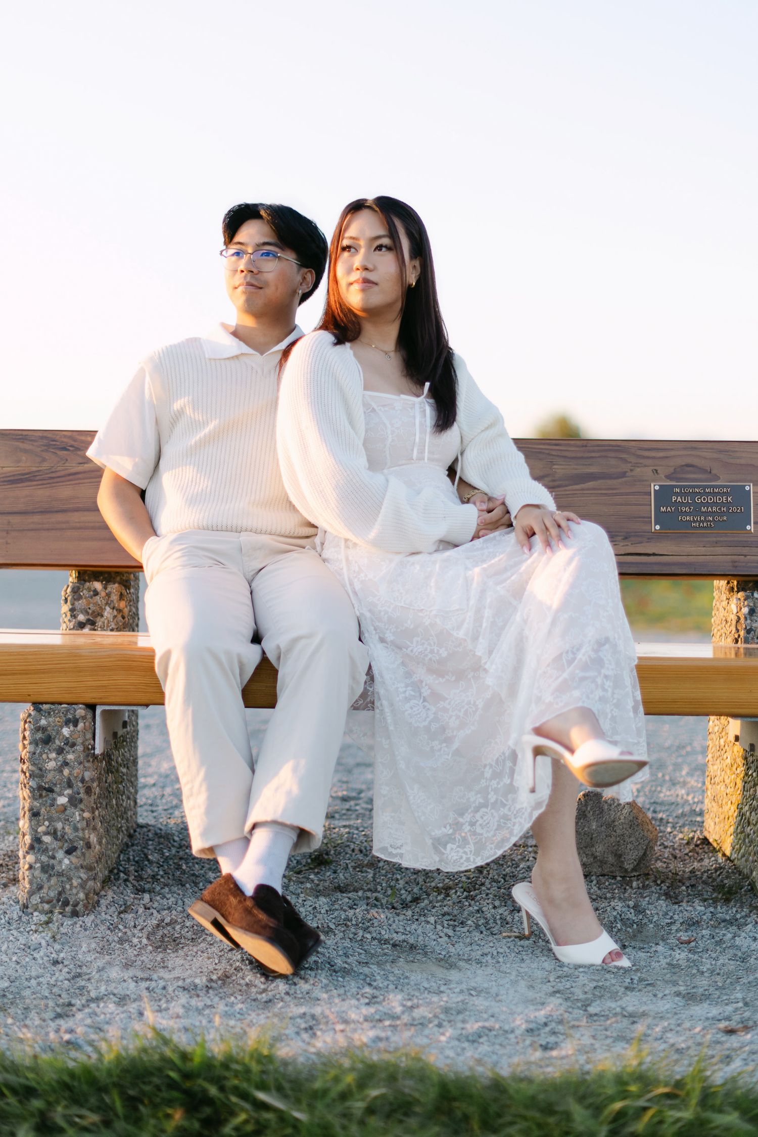 A couple in matching cream outfits sits close together on a wooden park bench at golden hour, gazing into the distance.