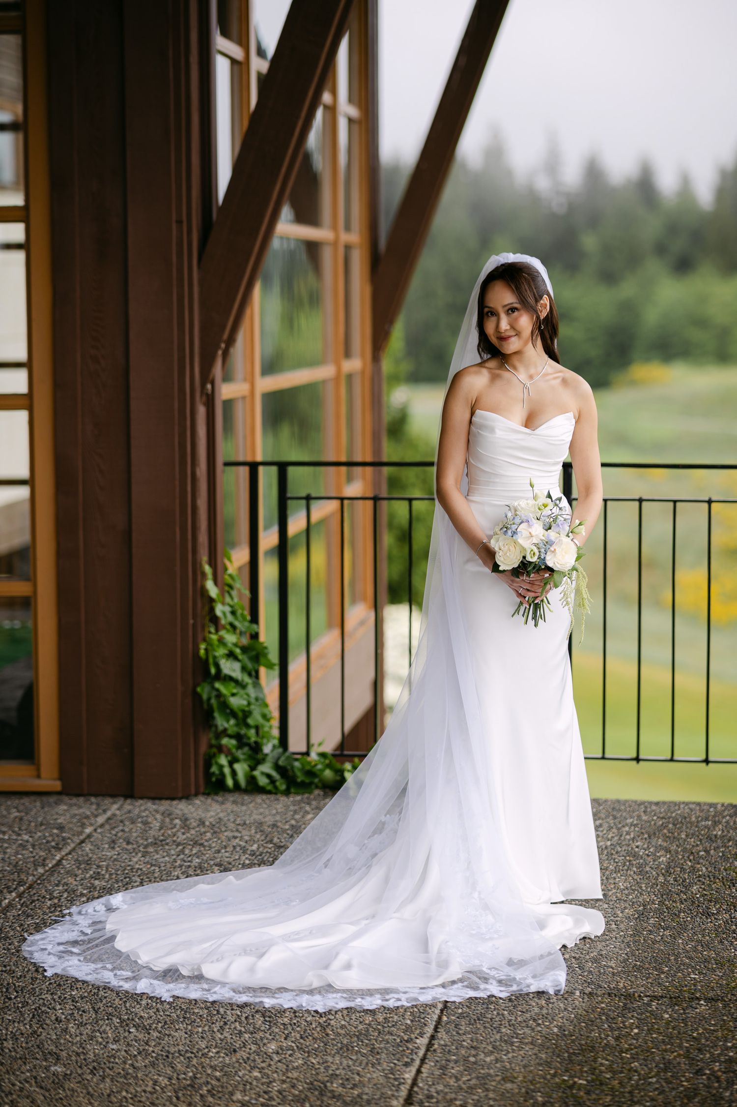 Bride in elegant strapless fitted white wedding dress with long train holding white bouquet outdoors.