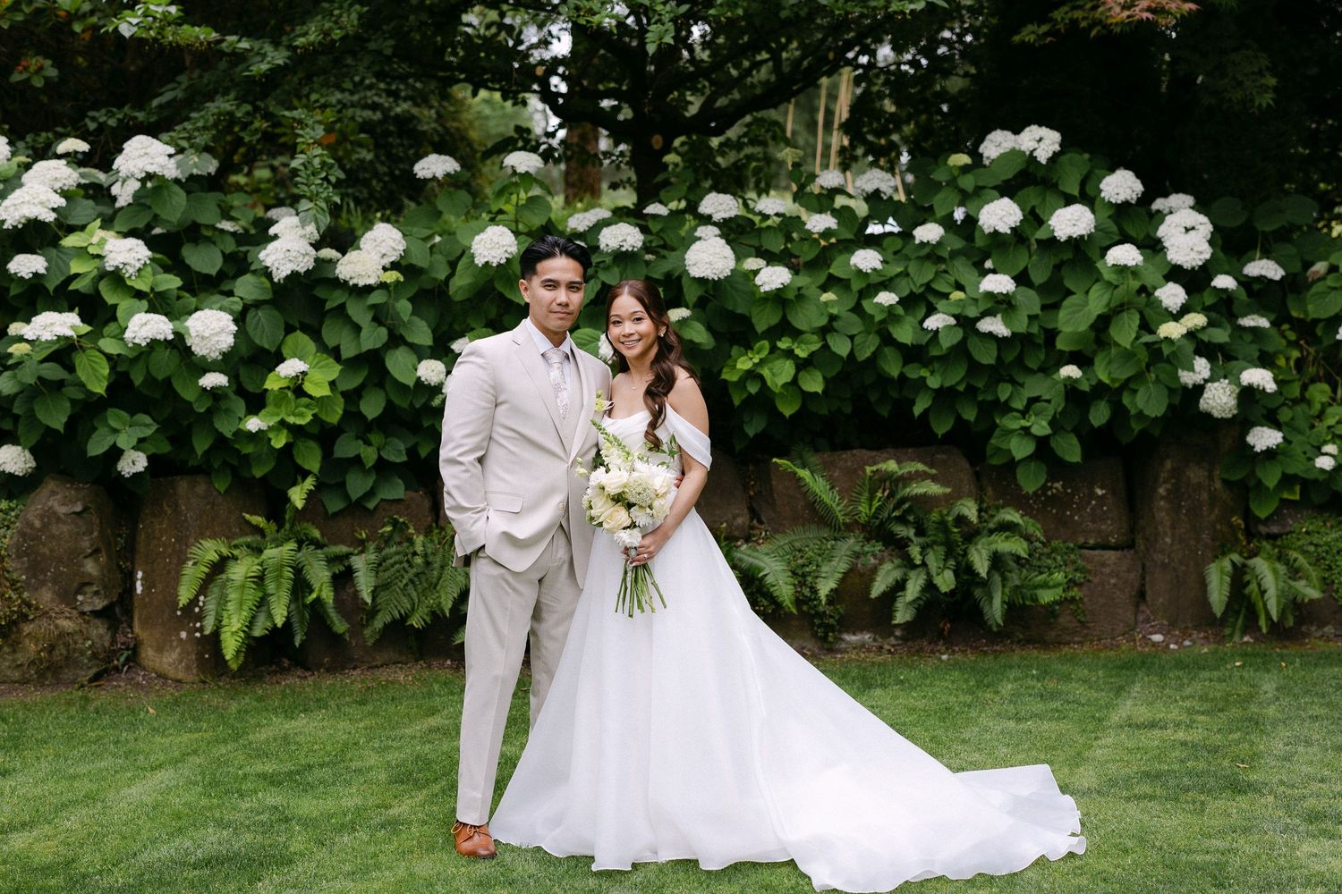 Bride and groom pose together in front of lush white hydrangeas in a garden wedding portrait.