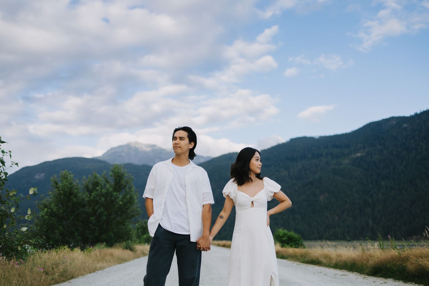 A couple holds hands on a country road, the woman in a white dress, with mountains and a cloudy sky behind them.
