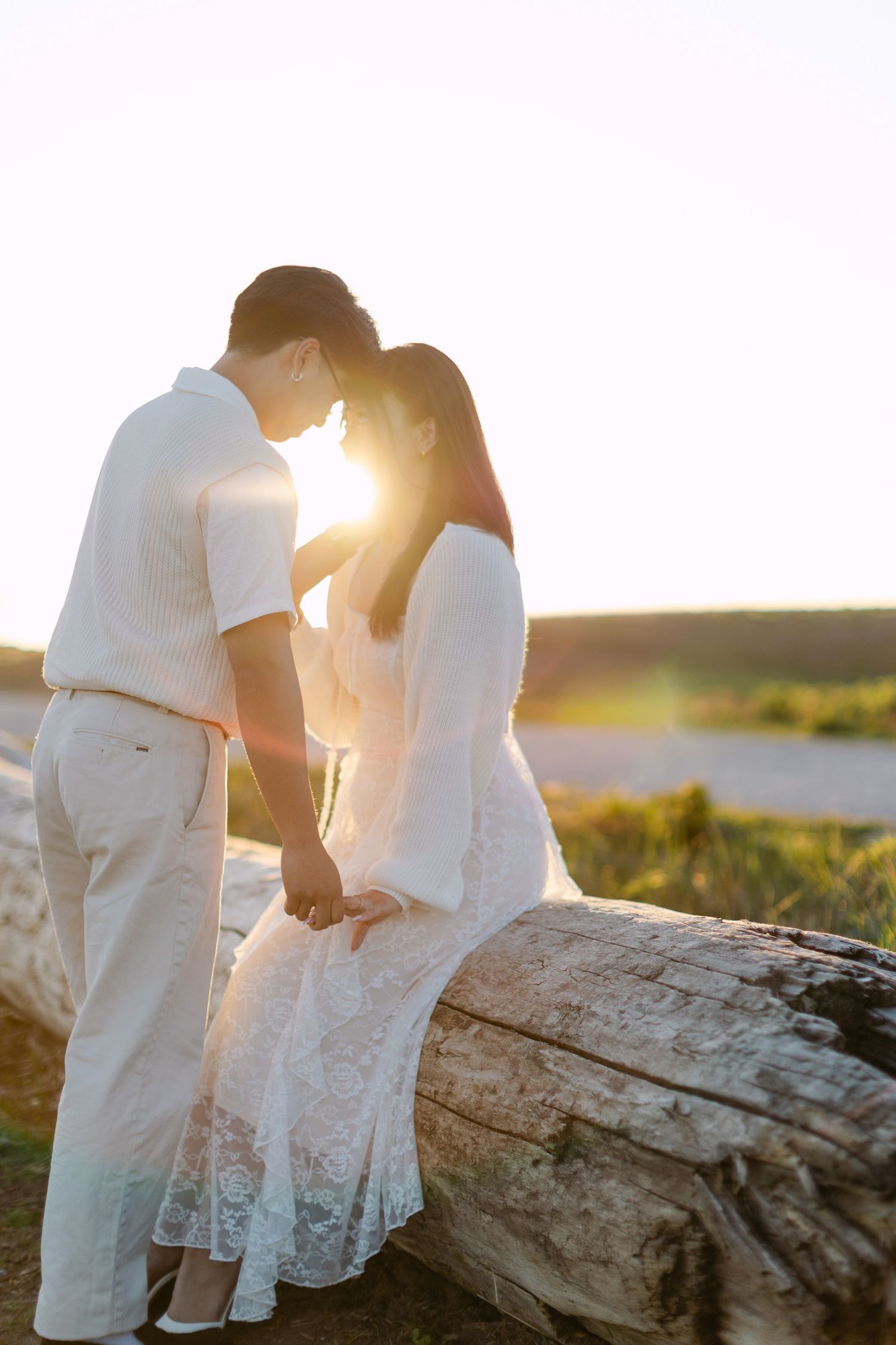 Couple touches foreheads and holds hands beside a weathered driftwood log as the sun glows between them.