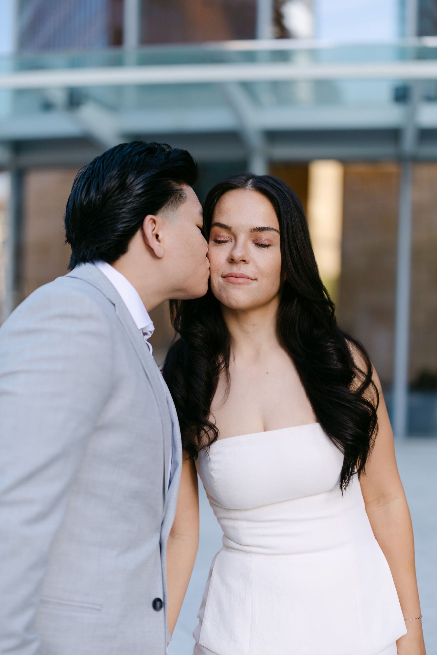 A man in a light gray suit kisses the cheek of a smiling woman in a white dress during an engagement photo session.