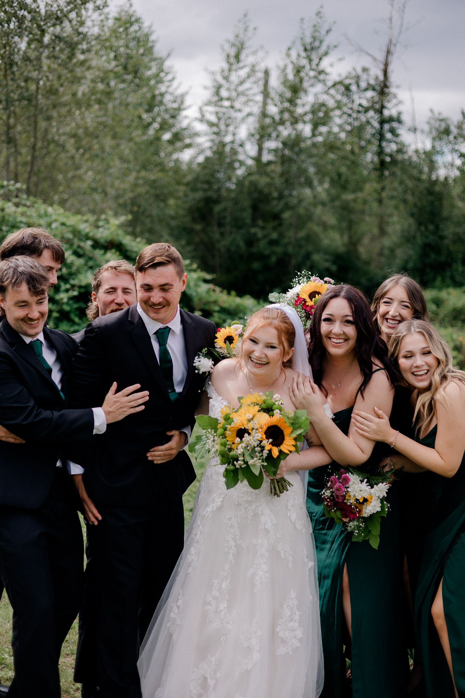 Wedding party in formal attire laughs and embraces outdoors with bridesmaids in dark dresses and groomsmen in black suits.