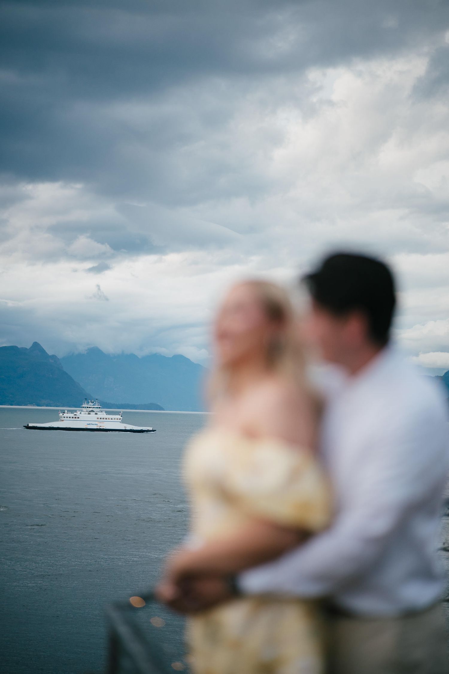 A couple overlooks a scenic ocean view with mountains and a ferry boat in the distance under moody cloudy skies.