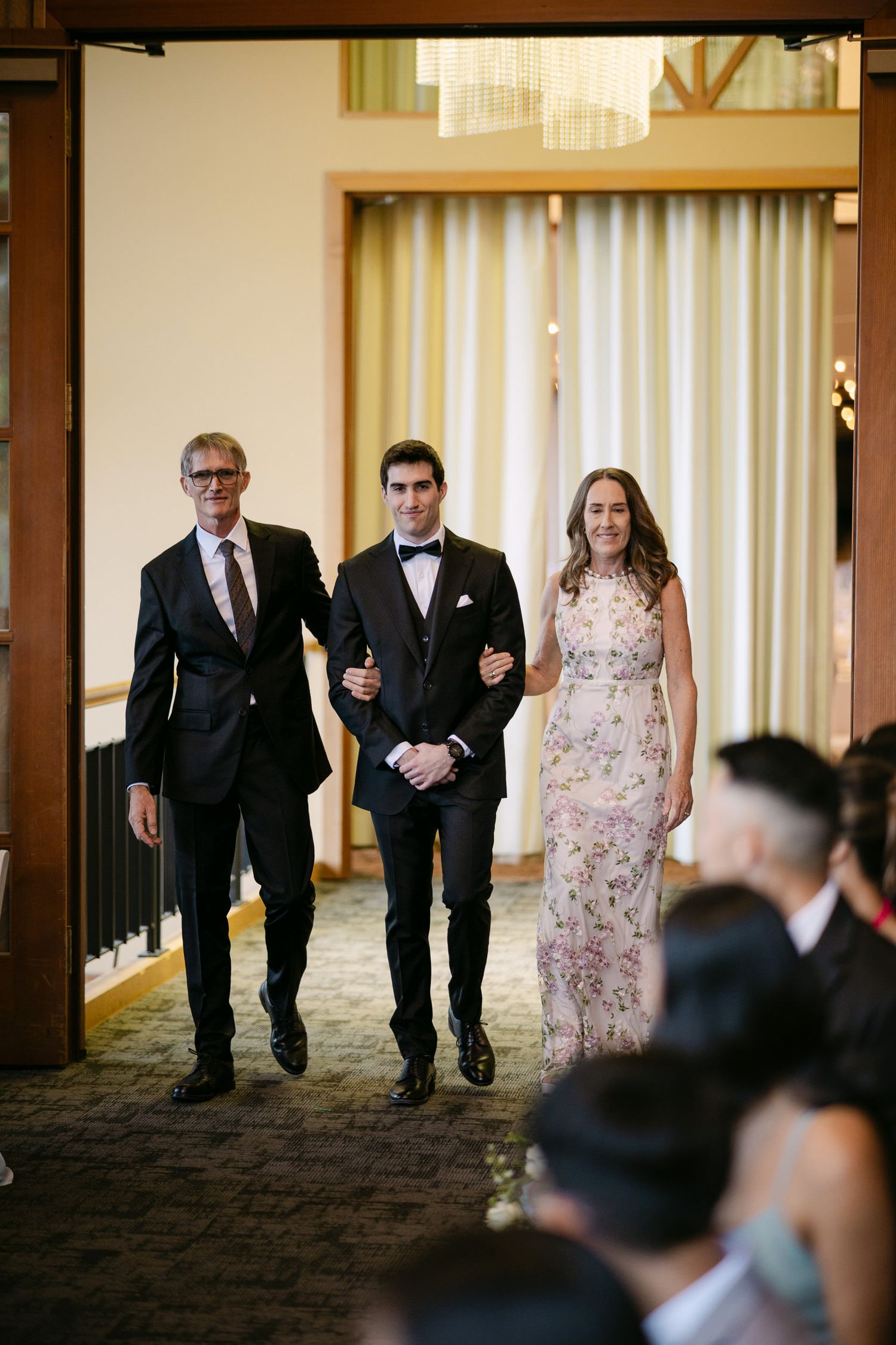 Groom walks down wedding aisle escorted by parents, wearing tuxedo, father in suit, mother in floral gown.