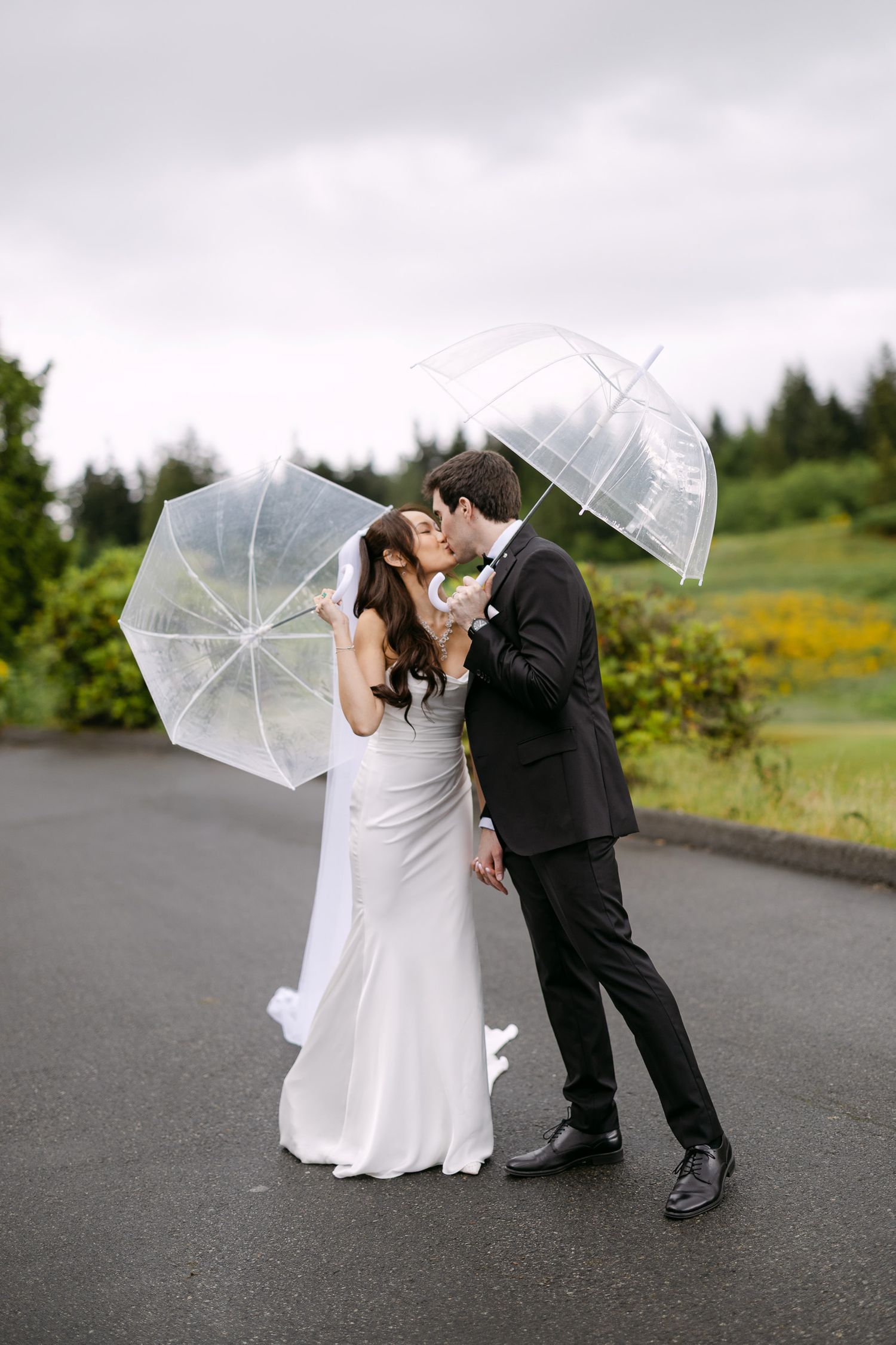 Bride and groom kissing in the rain under clear umbrellas on their wedding day.