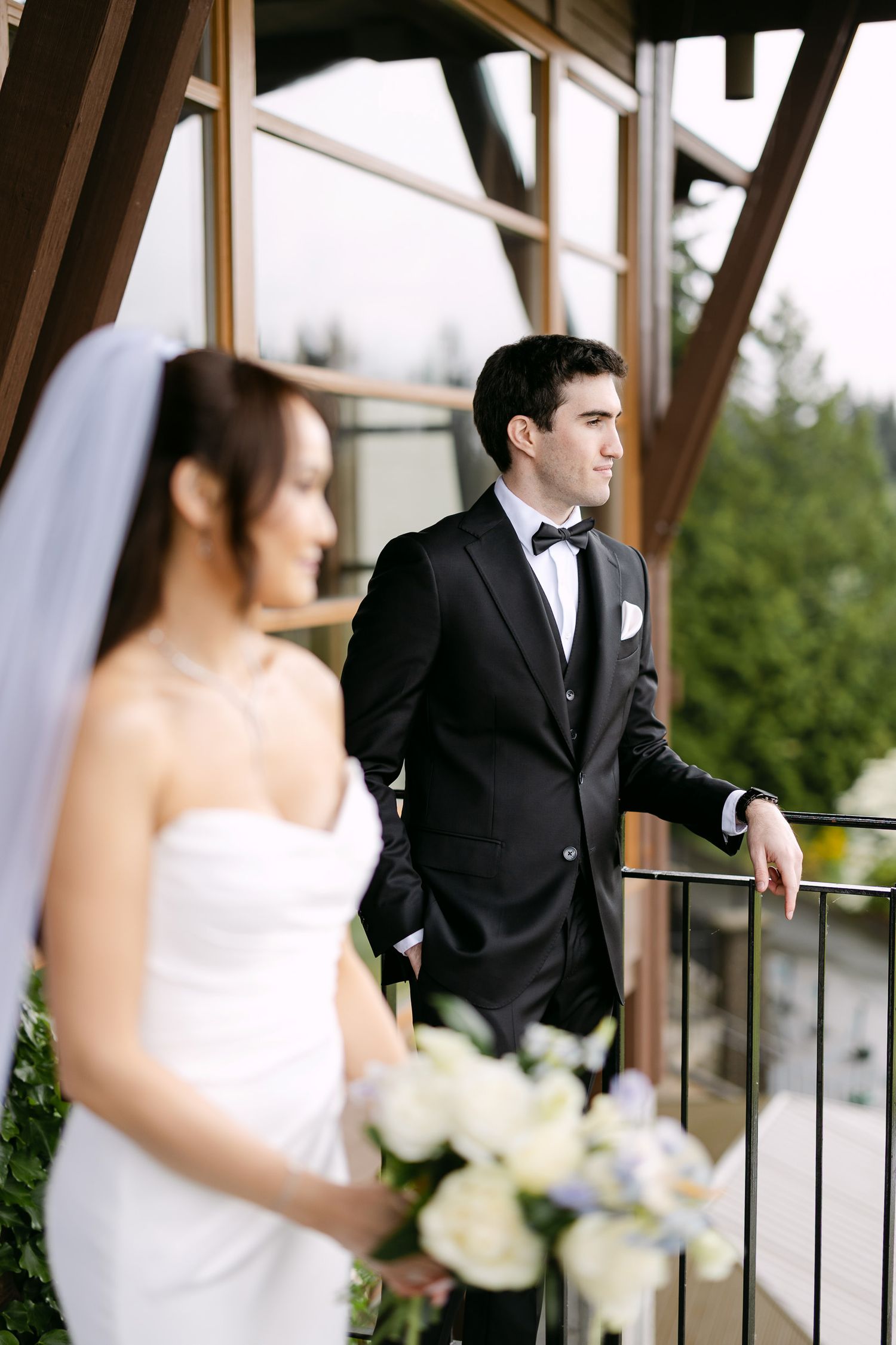 Bride in white strapless gown with veil and groom in black tuxedo stand on balcony with lush greenery backdrop.