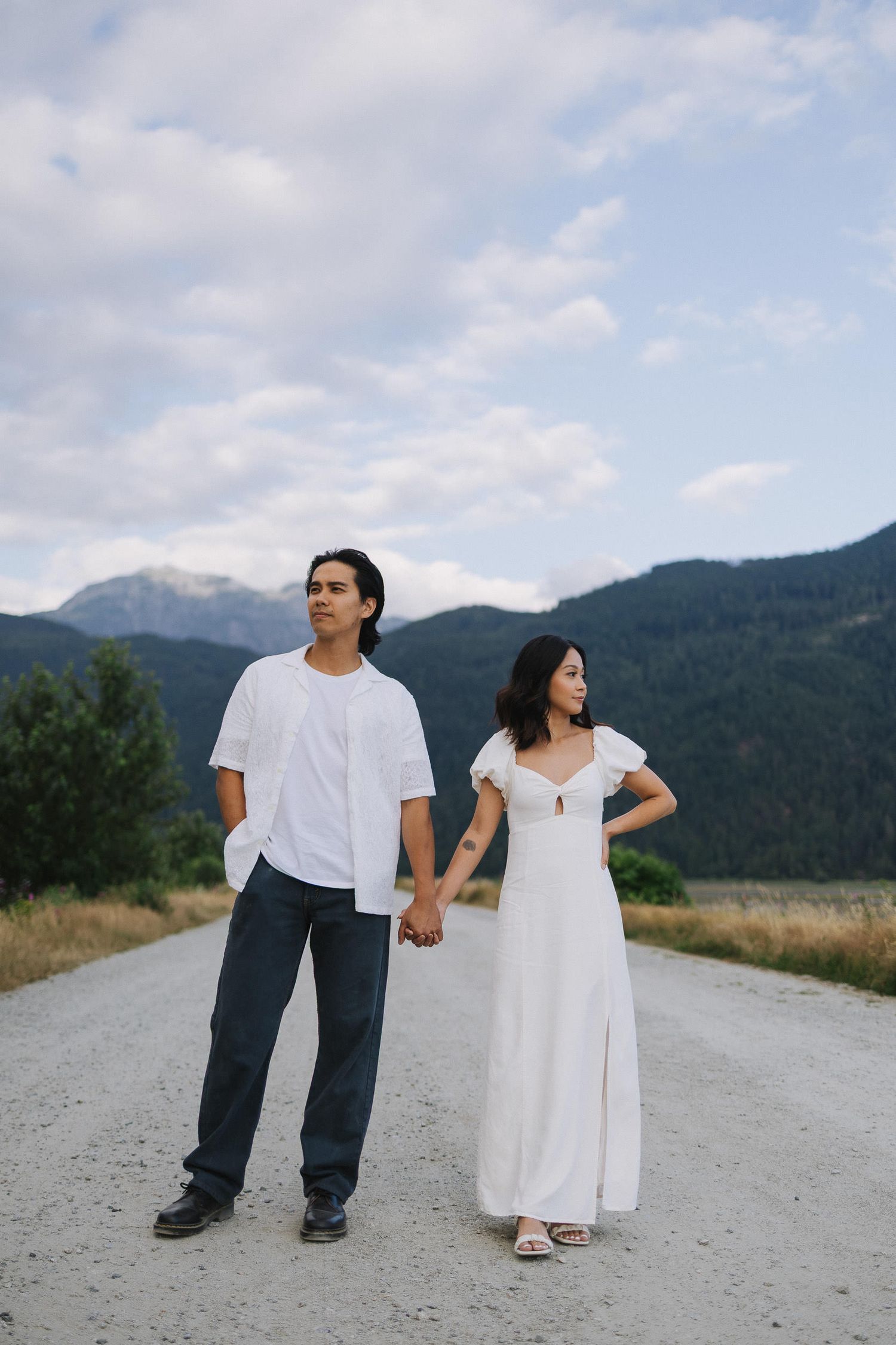 A couple in white and denim stands on a mountain path holding hands against a scenic mountain backdrop.