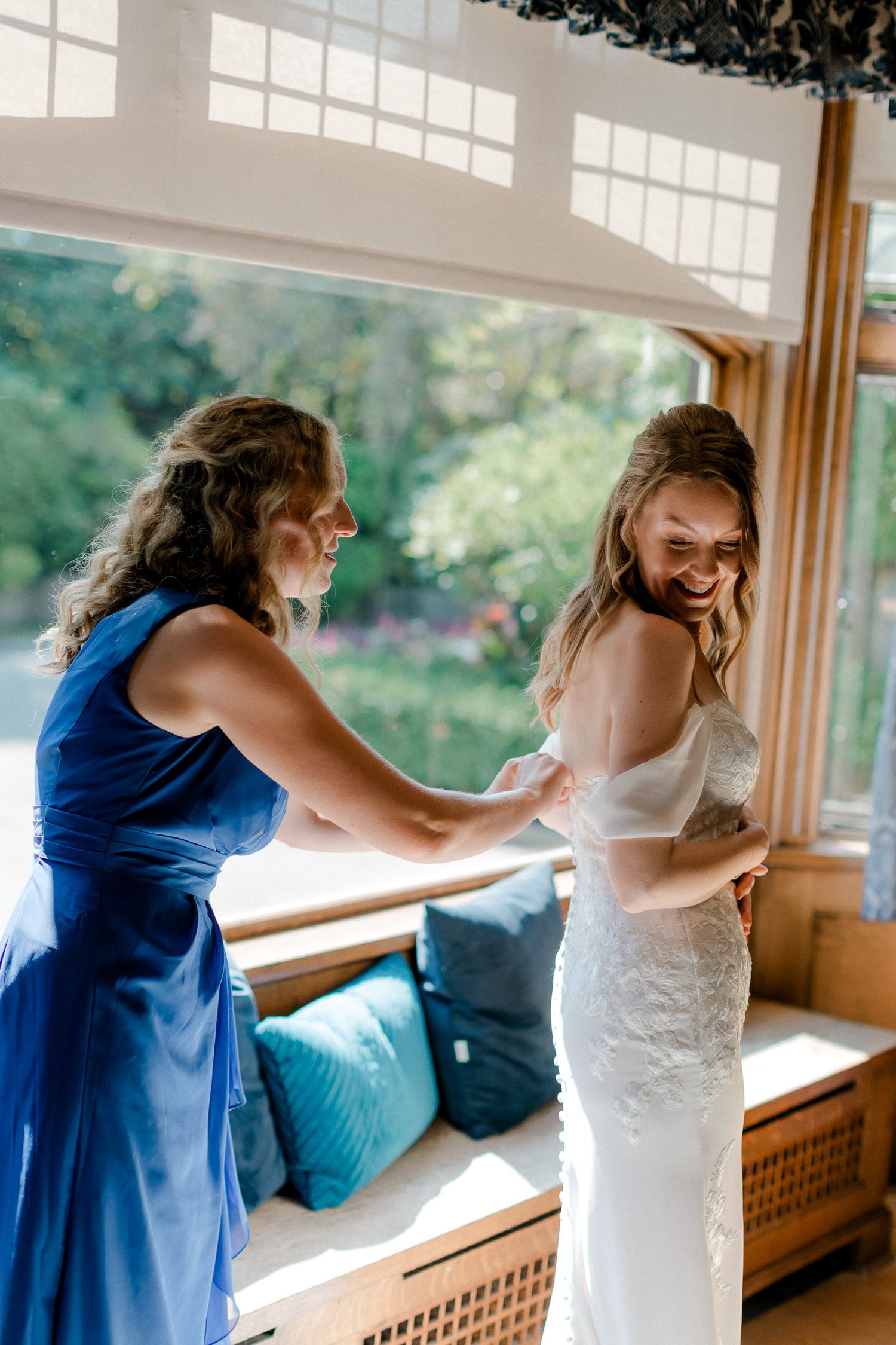 Two women share a joyful moment in a sunlit room as one helps adjust the other's white lace wedding dress.