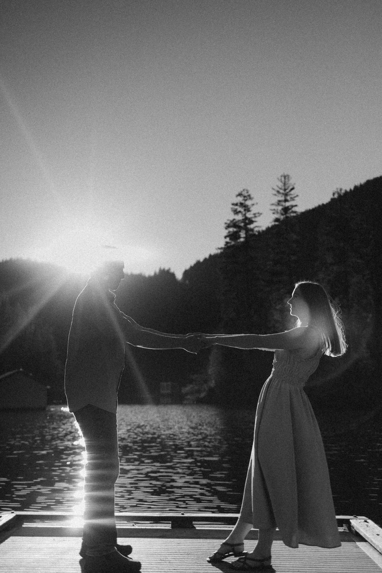 A silhouetted couple holds hands on a dock at sunset with mountains and trees in the background.