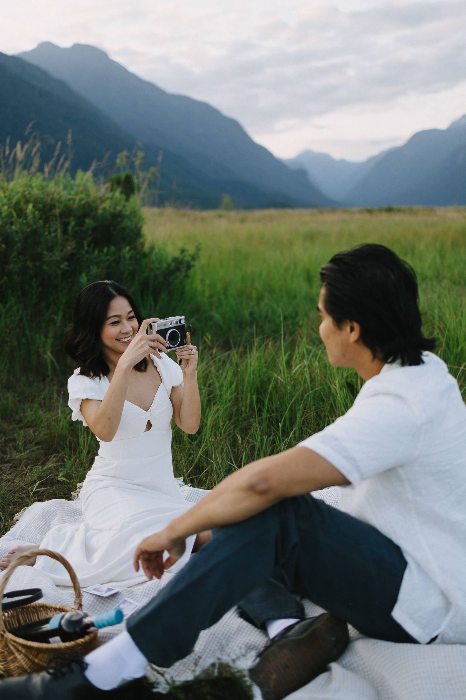 Two people in white shirts sit in a grassy meadow with mountains in the background taking photos together.