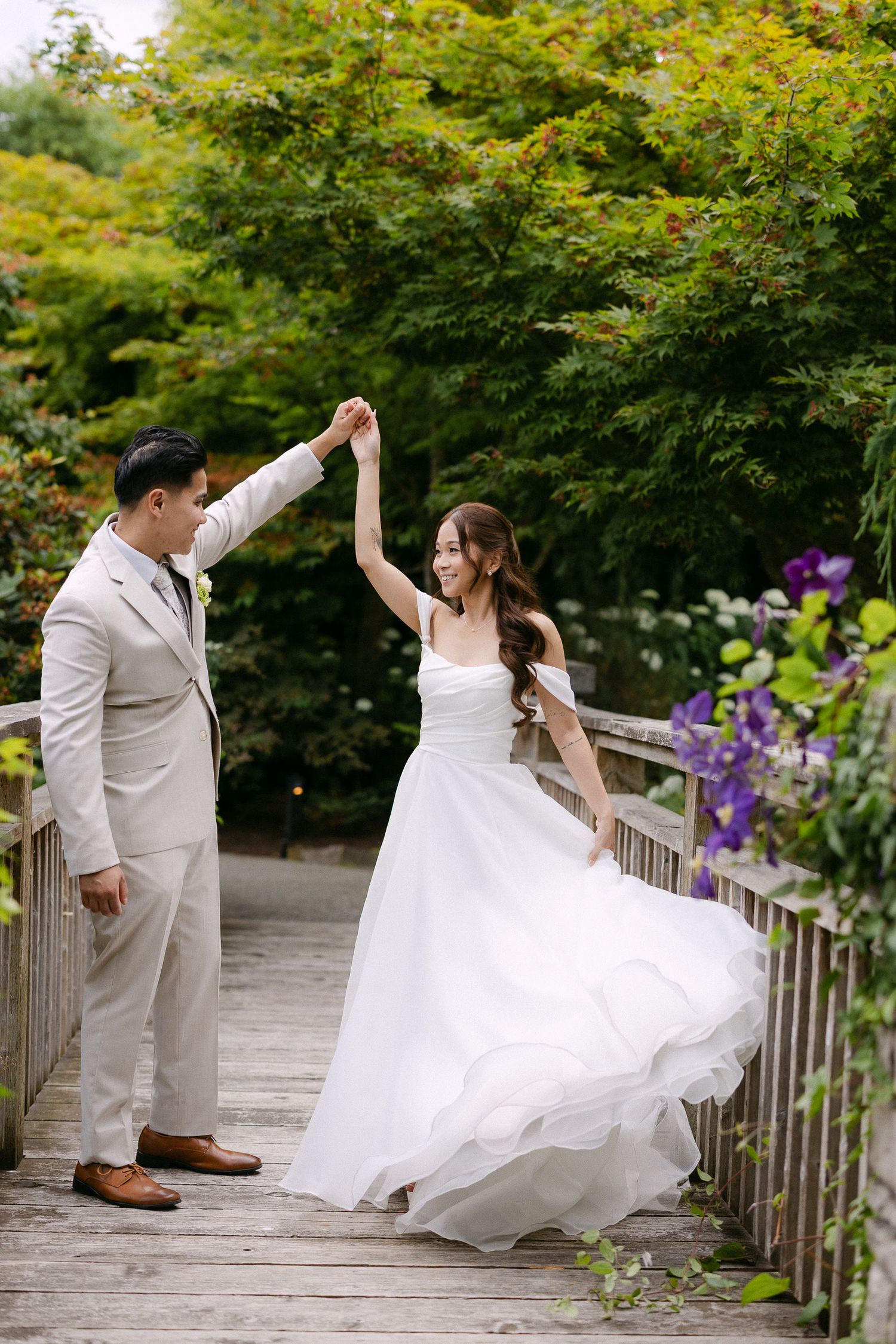 A couple dances on a wooden bridge, groom in beige suit twirling bride in flowing white wedding gown amid lush greenery.